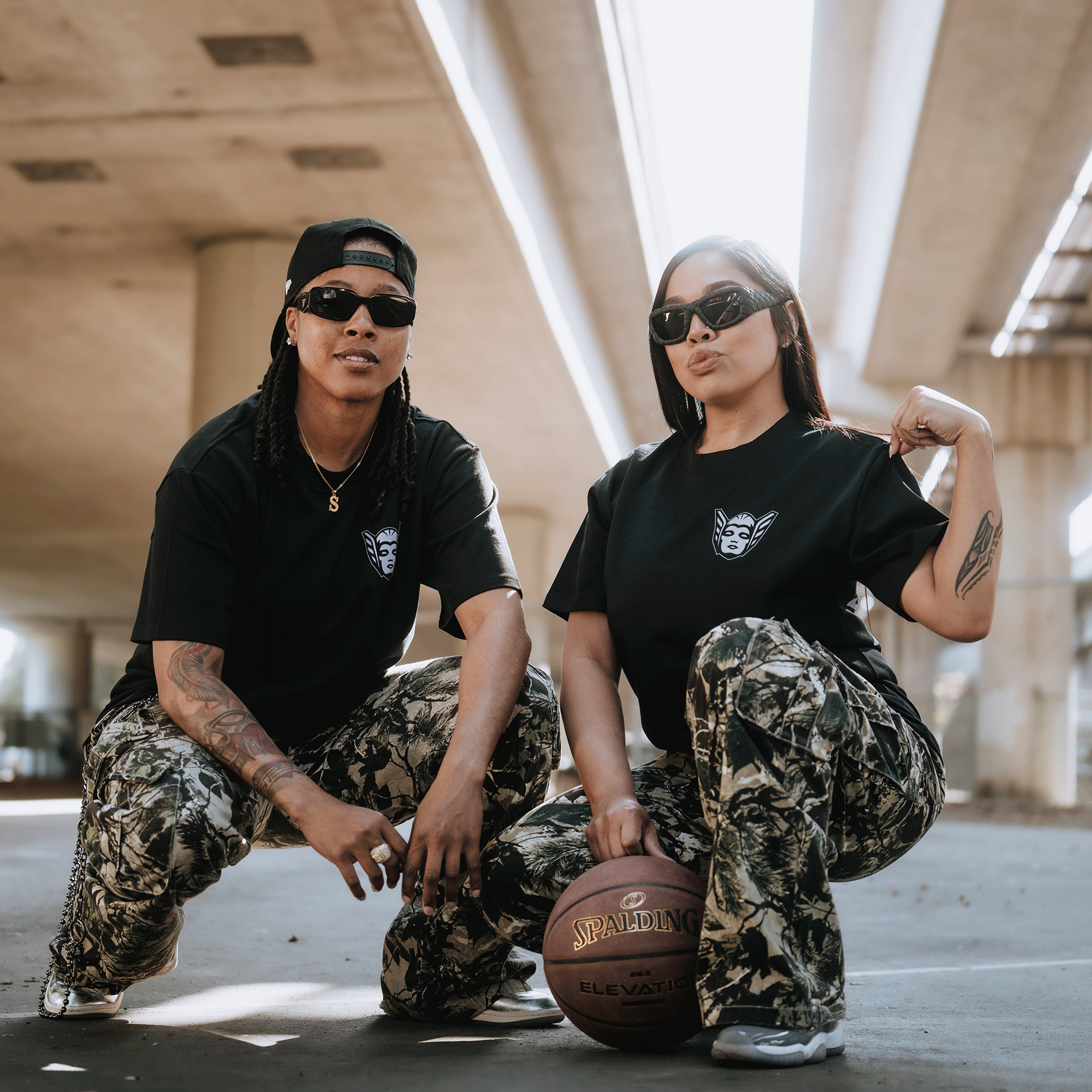 Two women pose under a sunlit overpass in GSV’s Golden State Valkyries Renowned Heavy Tees and camo pants—one kneels with a basketball while the other flexes, radiating team energy in an urban backdrop.