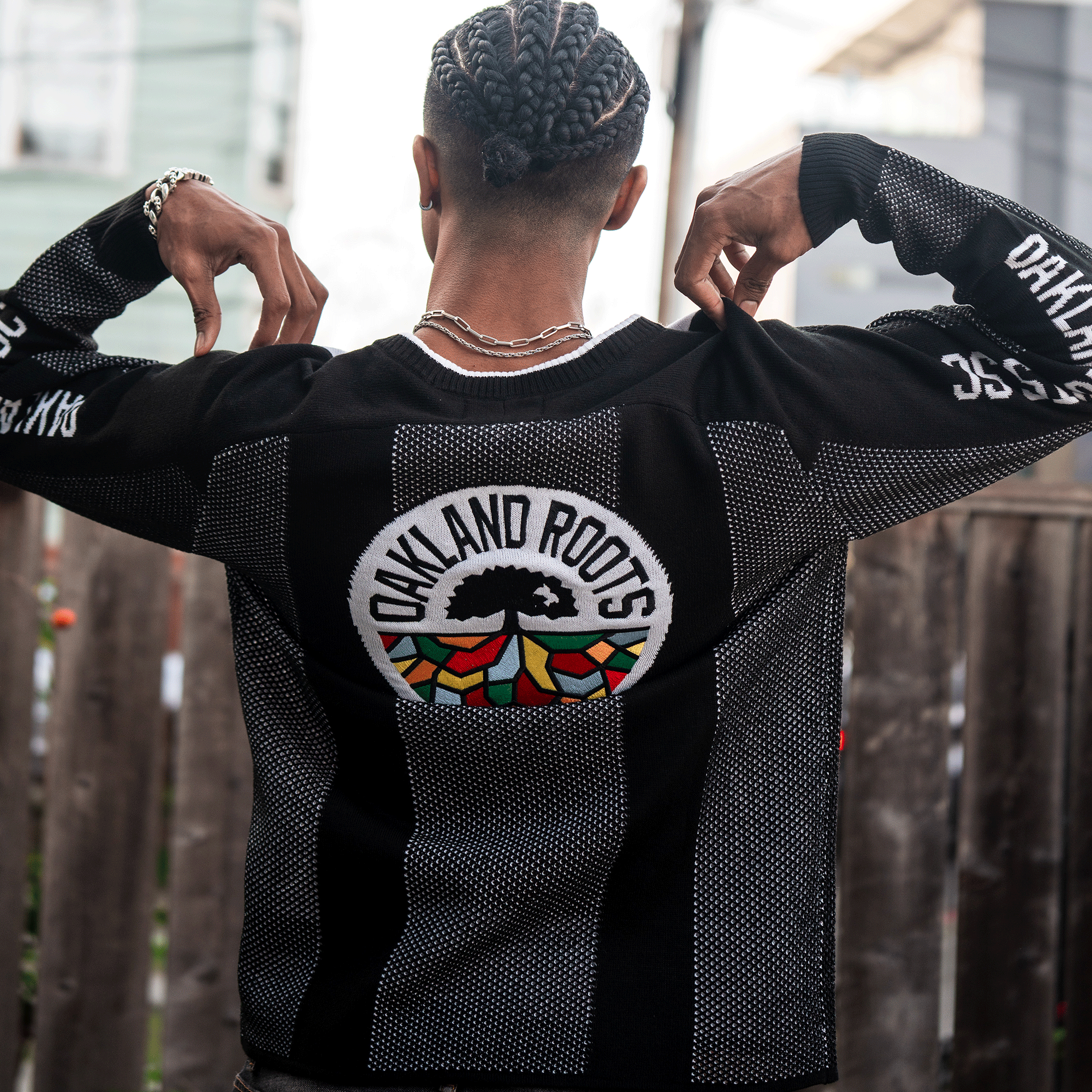 A person with braided hair lifts the collar of the Oakland Roots SC Clubhouse Sweater, showing off its large rainbow tree logo and “Oakland Roots” sleeves. A fence and houses are visible in the background. Brand: Oakland Roots SC.