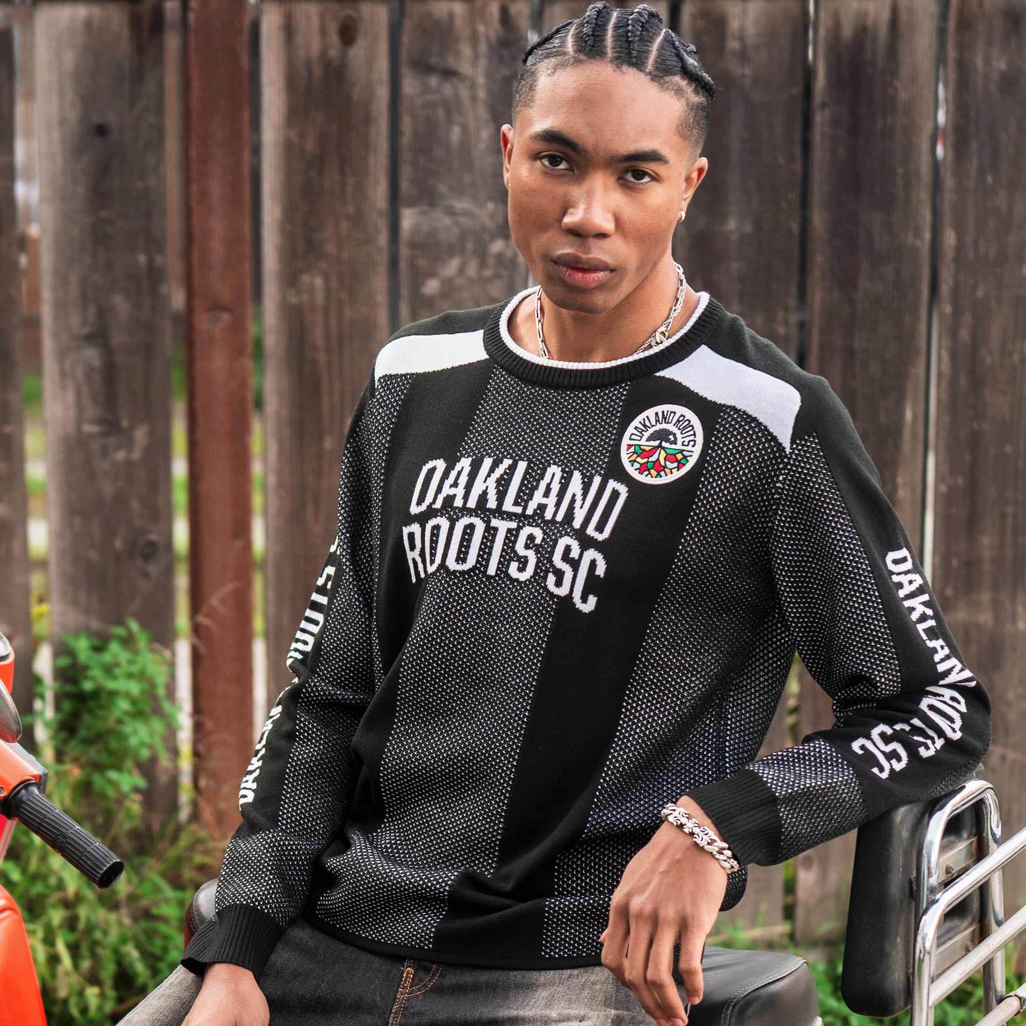 A young man with braided hair sits on a red bike by a wooden fence, wearing the Oakland Roots SC Clubhouse Sweater by Oakland Roots SC in unisex sizing. He faces the camera with a neutral expression, accessorized with a silver chain and bracelet.