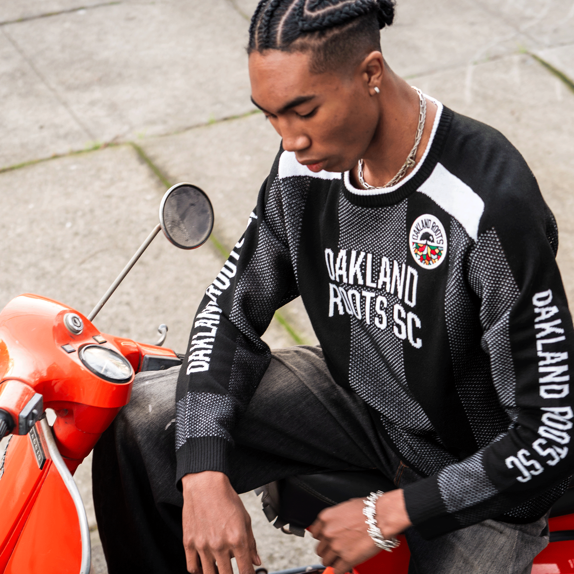 A young man with braided hair sits on an orange scooter, wearing a black Oakland Roots SC Clubhouse Sweater (unisex), dark jeans, a necklace, and earrings. The photo shows the sidewalk tiles and scooter handlebar from above.