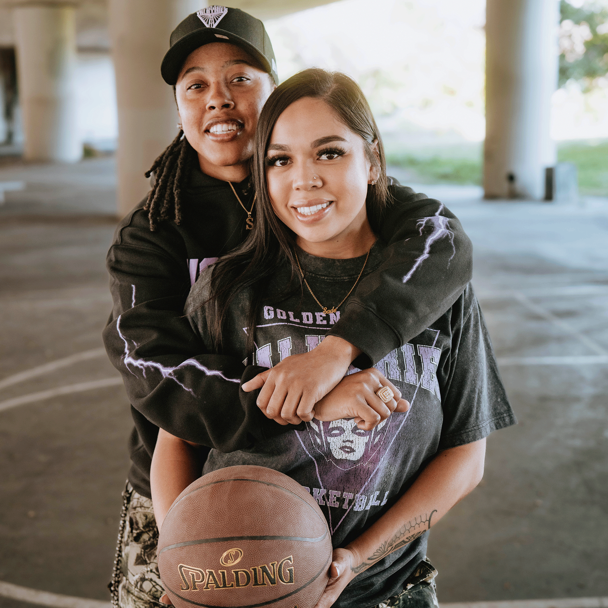Two people smile on a basketball court under an overpass. One wears a GSV Golden State Valkyries Violet Lightning Hoodie, standing behind with arms around the other, who holds a Spalding basketball as daylight streams in.