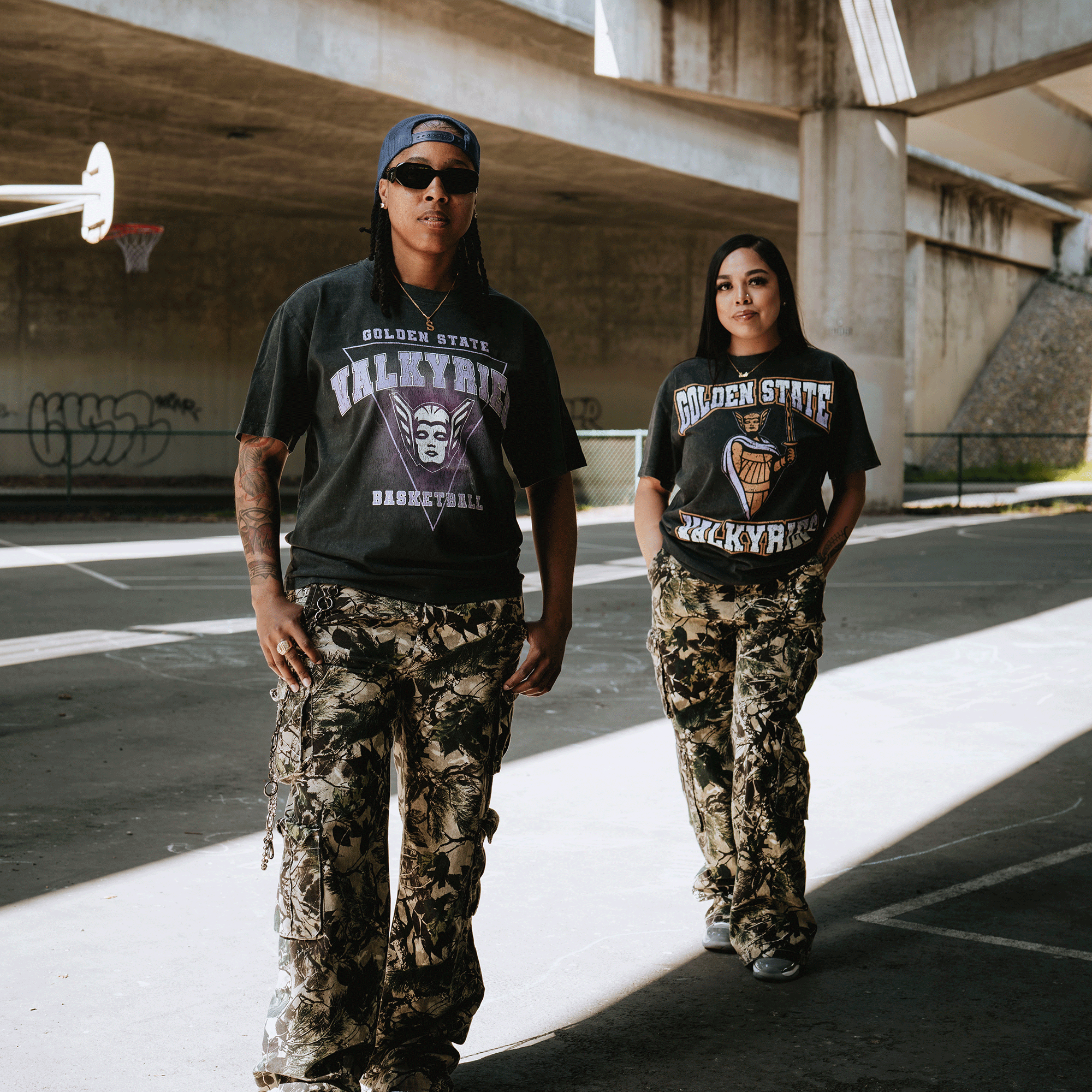 Two people stand confidently on a basketball court under an overpass, both wearing GSV Golden State Valkyries Surge Stone Wash Heavy Tees and camouflage pants, as sunlight casts sharp shadows on their standout style.