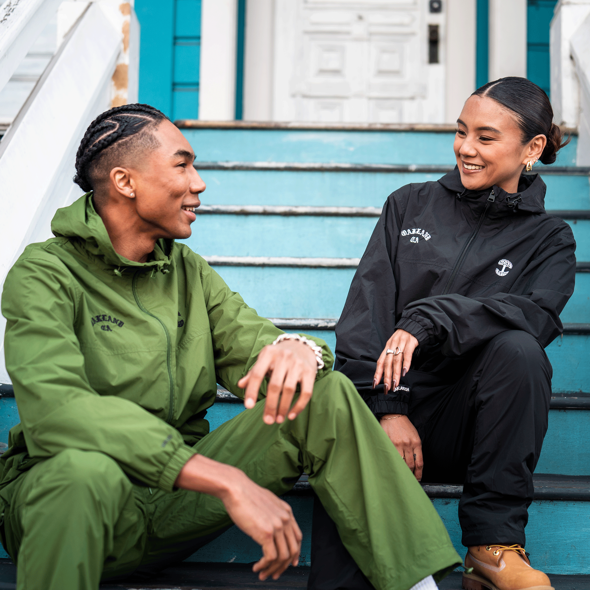 Two people sit on blue wooden steps, smiling at each other. The person on the left wears the Oaklandish Susurrus Track Bottom in green with an adjustable waistband and braided hair; the other wears a black jacket and tan boots. A white door and railing are behind them.