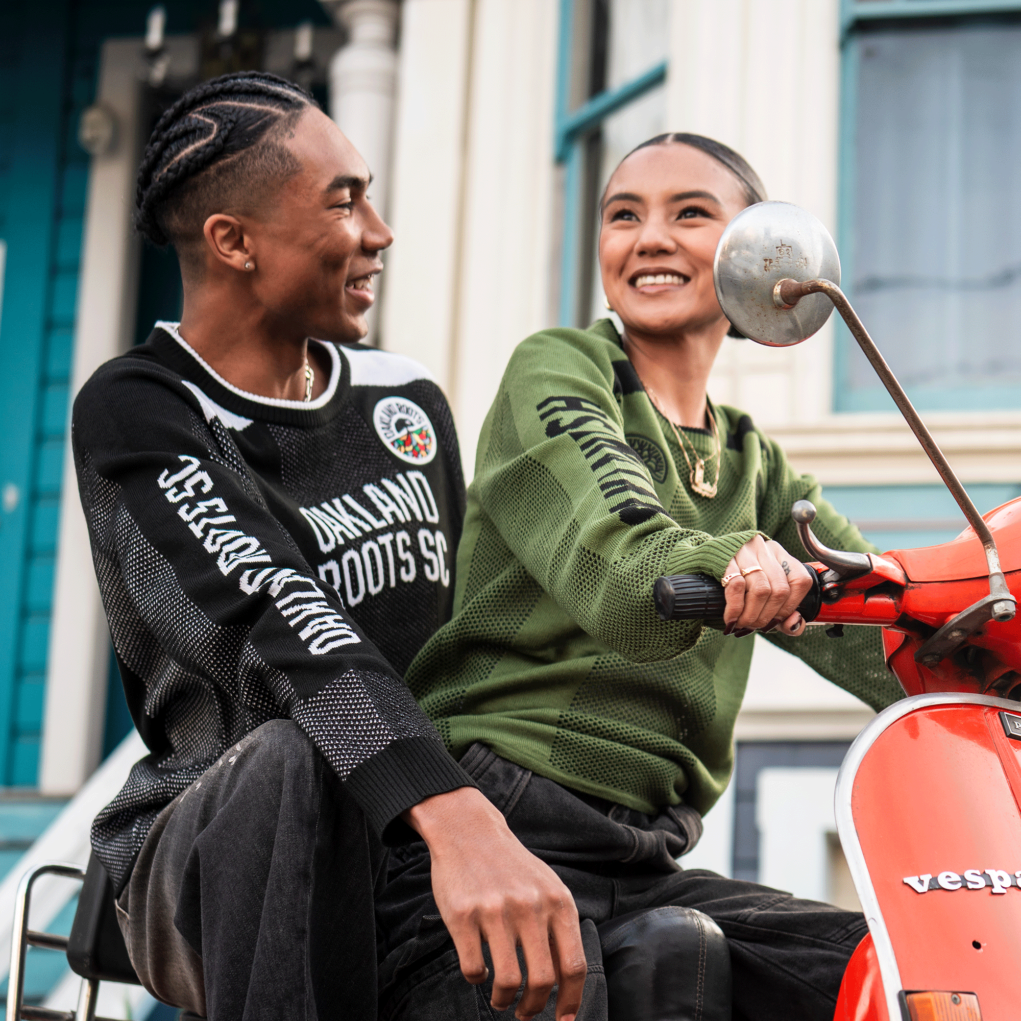 A smiling woman in a green knit sweater sits on an orange Vespa, holding the handlebars, while a man in a black Oaklandish Terrace Sweater by Oaklandish sits behind her. They are outside in front of colorful buildings, looking at each other.