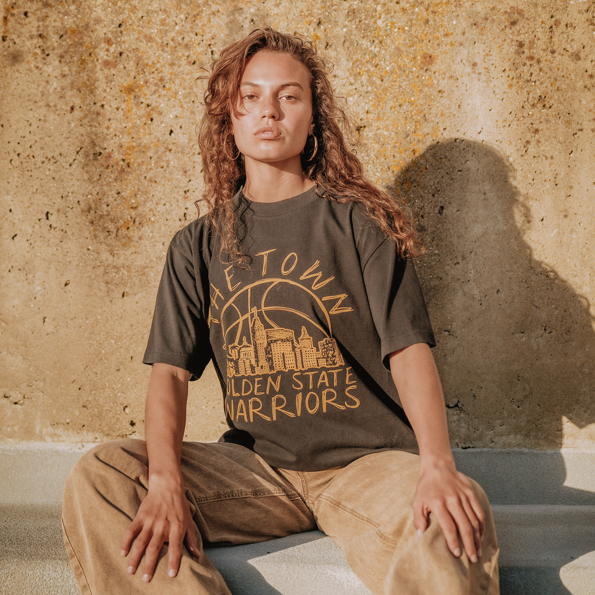 A woman with wavy brown hair sits on concrete steps in sunlight, wearing a dark GSW Town Doodle Tee and tan pants. A city skyline graphic is visible on her shirt as she gazes confidently at the camera, her shadow cast boldly on the wall.