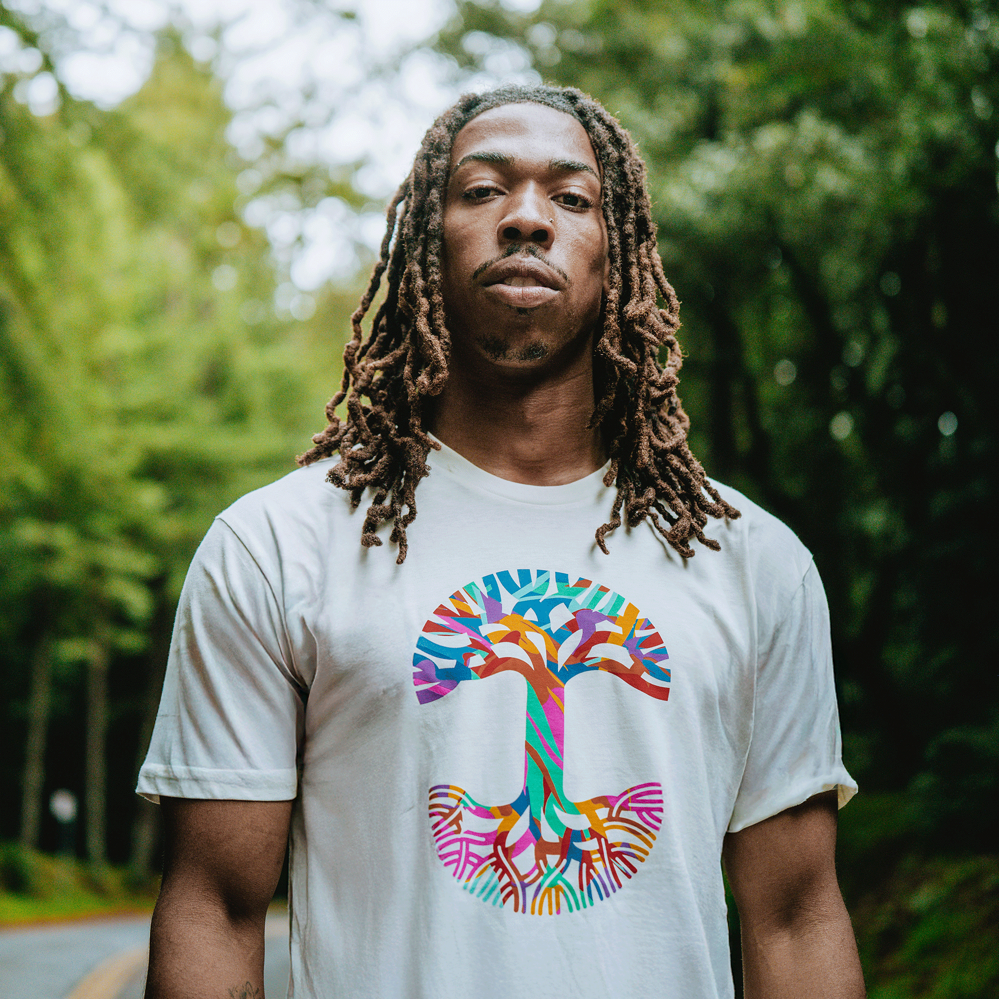 A man with long brown dreadlocks stands outdoors before green trees, wearing the Oaklandish Urban Tropics Tee, which features a vibrant abstract tree design inspired by multicultural influences. The background is blurred.