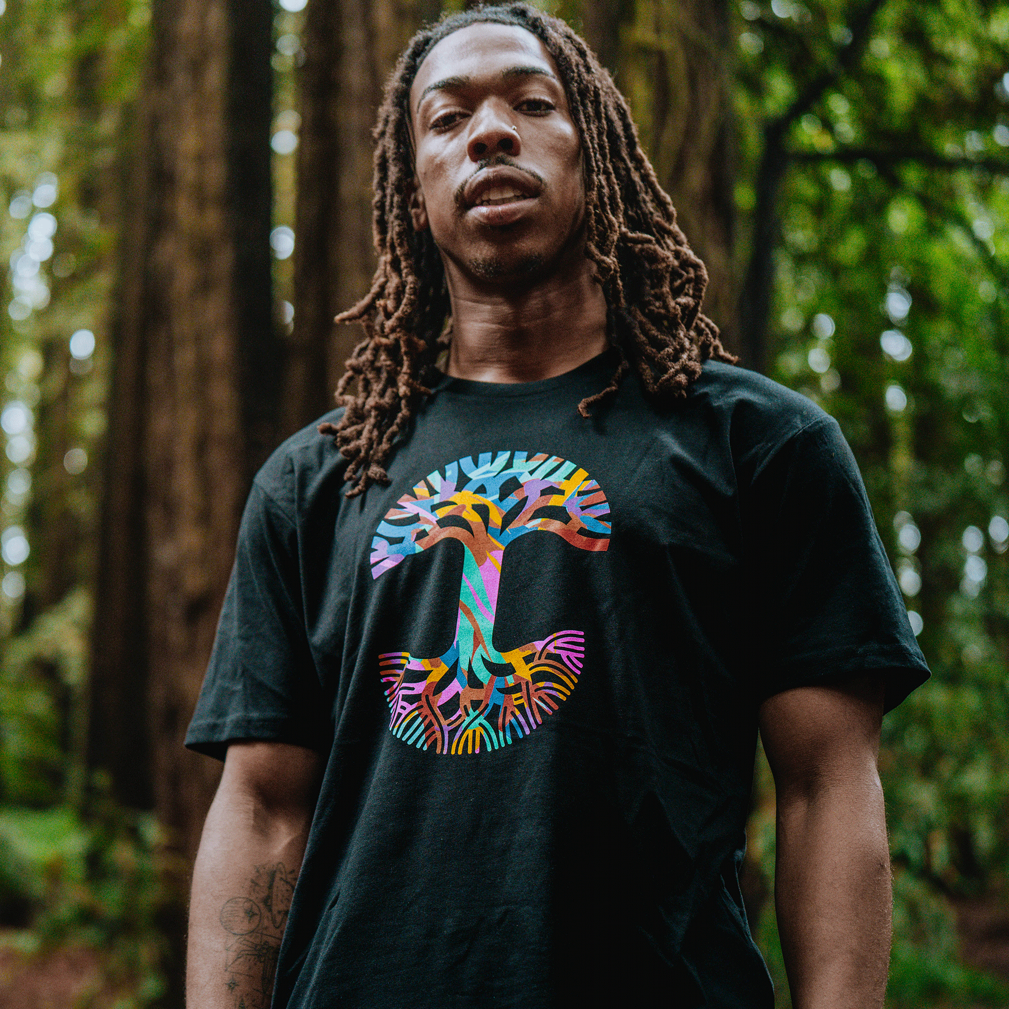 A man with long dreadlocks stands confidently in a lush forest, sporting the Urban Tropics Tee by Oaklandish, featuring a bold, colorful tree design that celebrates multicultural art amid vibrant green foliage.