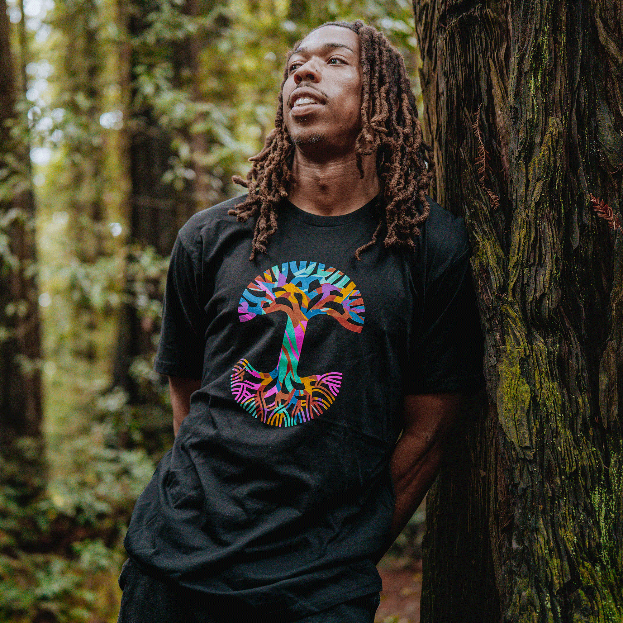 A man with long brown dreadlocks leans against a tree in a forest, wearing the Oaklandish Urban Tropics Tee, which features a large, colorful mushroom graphic. He's surrounded by green foliage with soft light filtering through the trees.