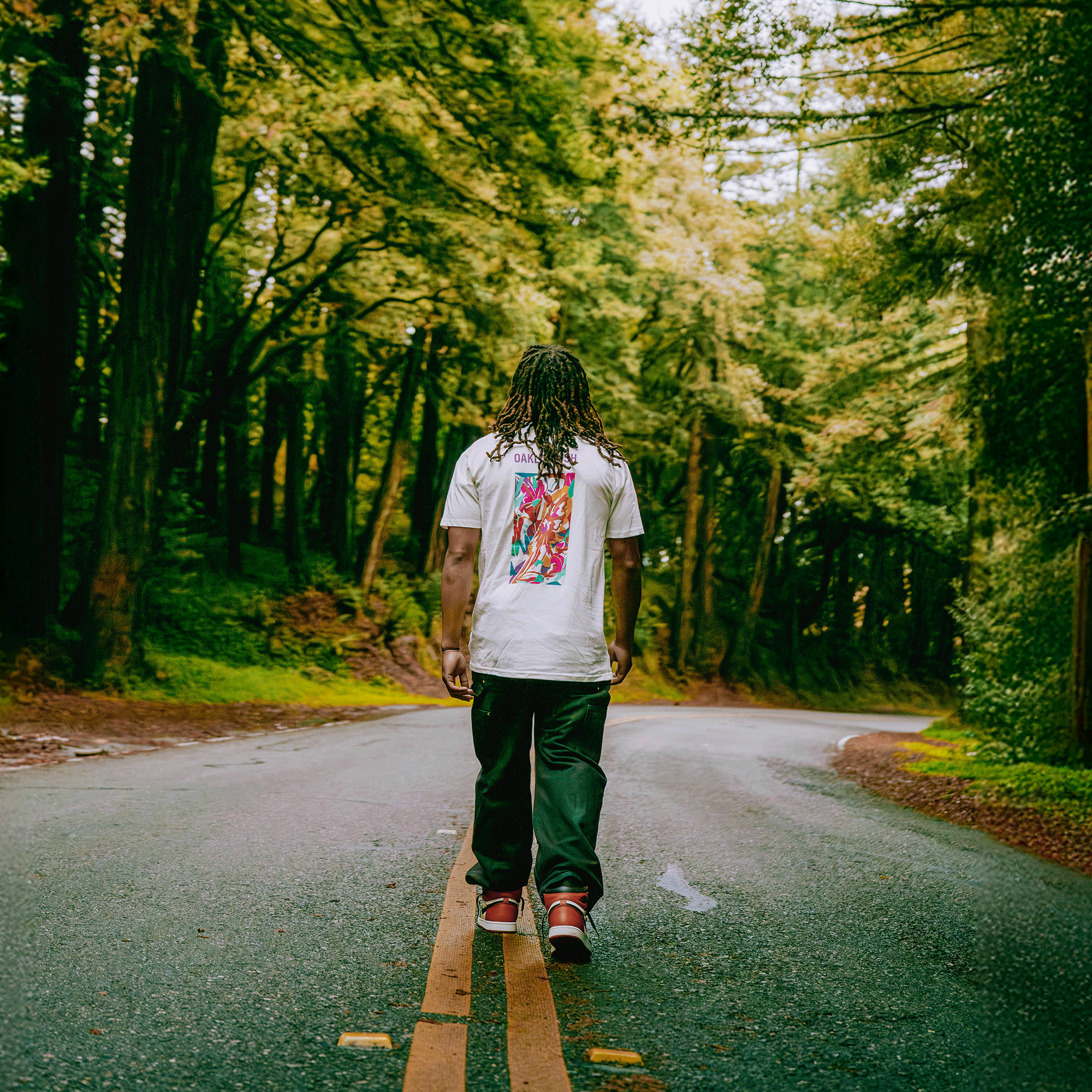A person with long, braided hair walks alone on a winding road in a lush forest, wearing the Oaklandish Urban Tropics Tee, dark jeans, and red sneakers, surrounded by towering trees and vibrant greenery.