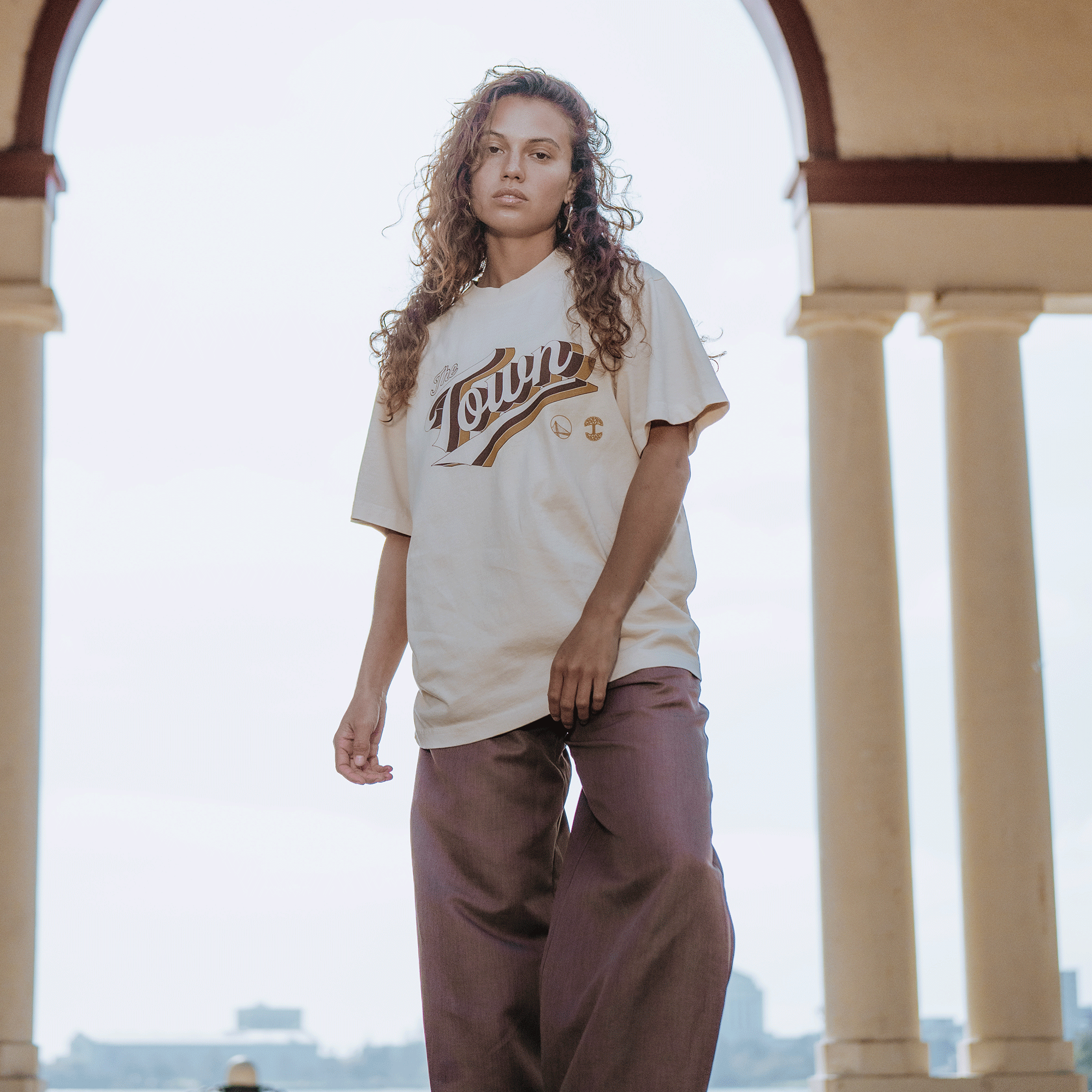 A woman with long curly hair stands under an archway, wearing the GSW Vintage Swash Tee with wide-legged brown pants. She gazes confidently at the camera, a cityscape blurred in the background.