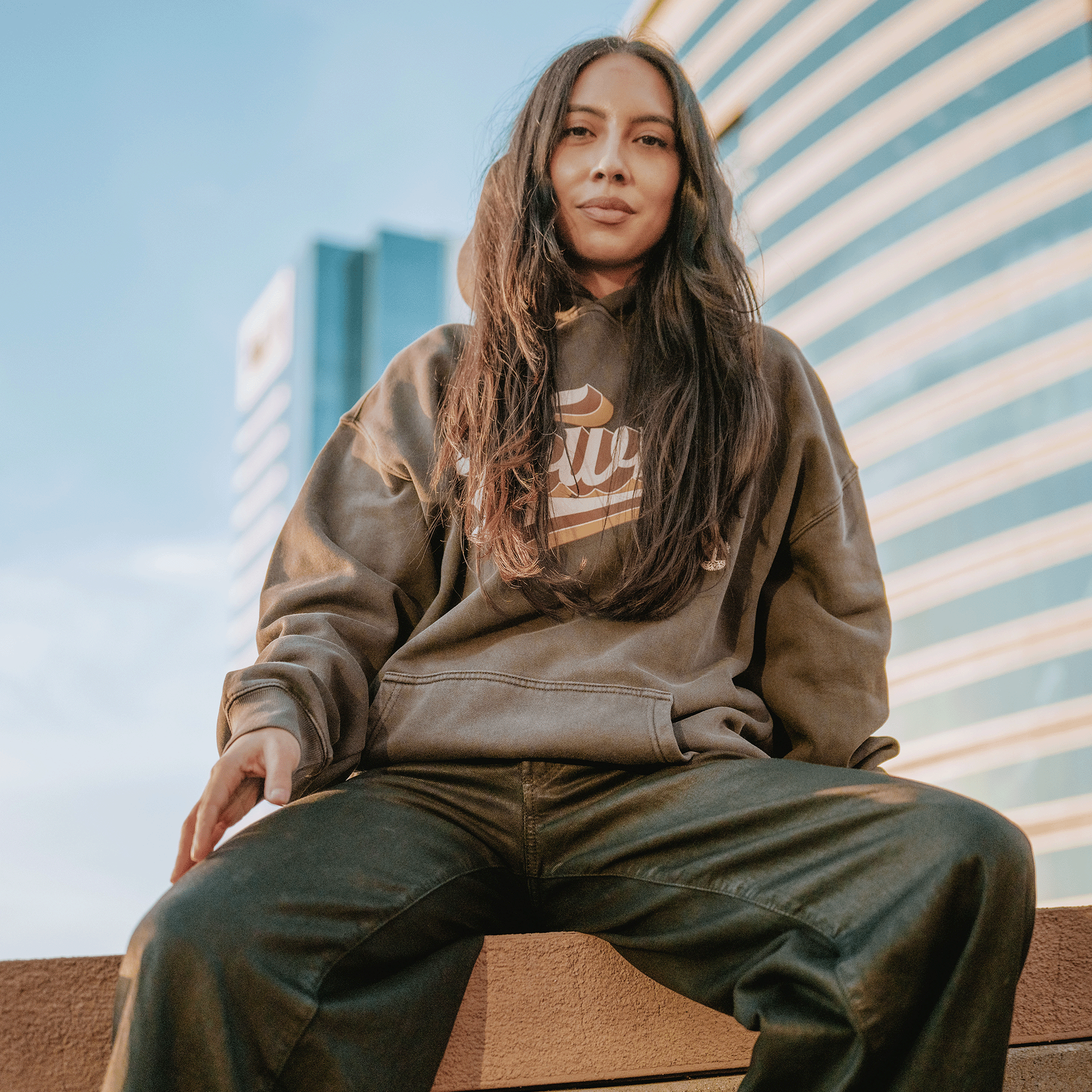 A woman with long brown hair sits on a ledge outdoors, confidently wearing the GSW Vintage Swash Hoodie. Sunlight warms her face as modern glass buildings form a striking backdrop.