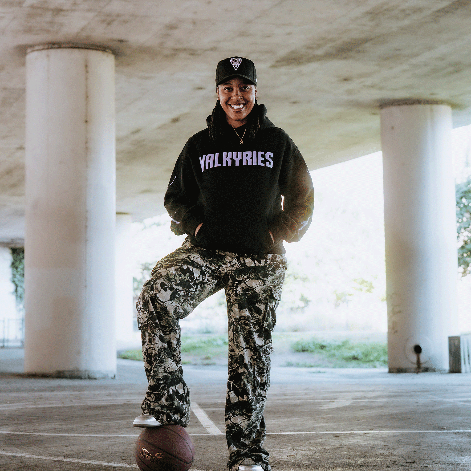 A person smiles under a bridge, wearing a black GSV Golden State Valkyries Violet Lightning Hoodie and camouflage pants with a black cap. They rest one foot on a basketball on concrete, with columns and greenery in the background.