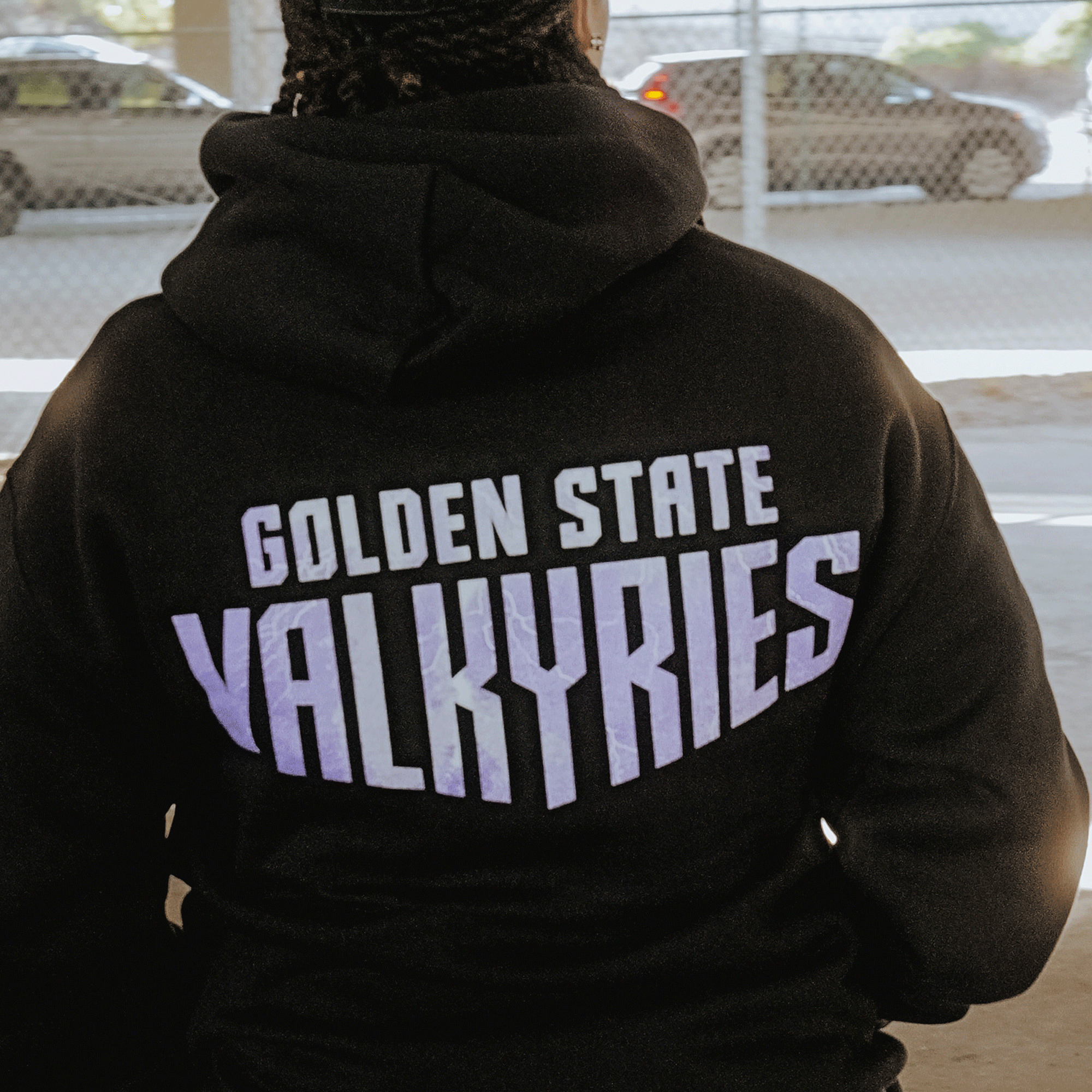 A person wears the GSV Golden State Valkyries Violet Lightning Hoodie, an officially licensed black mid-weight fleece pullover with "GOLDEN STATE VALKYRIES" in bold on the back, seen outdoors near a chain-link fence and blurred cars.
