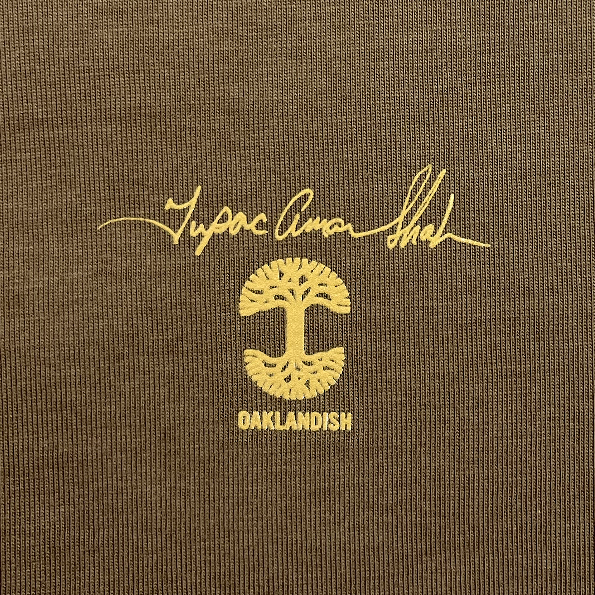 The "Against the World Tee" by Oaklandish features brown fabric adorned with a striking yellow "Oaklandish" logo. This hip-hop-inspired design includes a symmetrical tree with both branches and roots, surrounded by stylized text that reads "Home Grown Soul" above the tree and "OAKLANDISH" below it, set against a vividly textured fabric background.