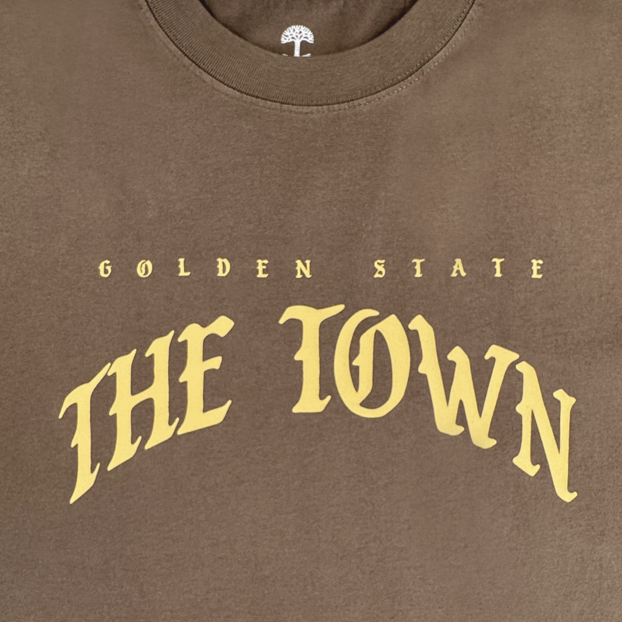 The GSW Arched Black-Letter Tee features “GOLDEN STATE” in small yellow letters above large, arched yellow text reading “THE TOWN.” A white tree logo is visible inside the collar. Part of the Town Collection.