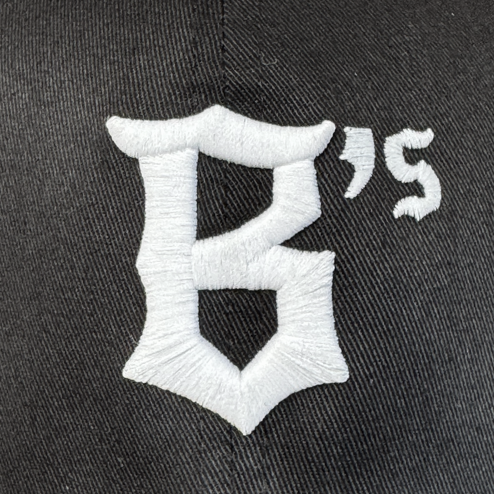 Close-up of black polyester fabric from the Oakland Ballers Flexfit Cap, featuring a large, ornate white "B" with a small white "'s" embroidered beside it—part of the Oakland Ballers logo—with visible stitching on the textured material.