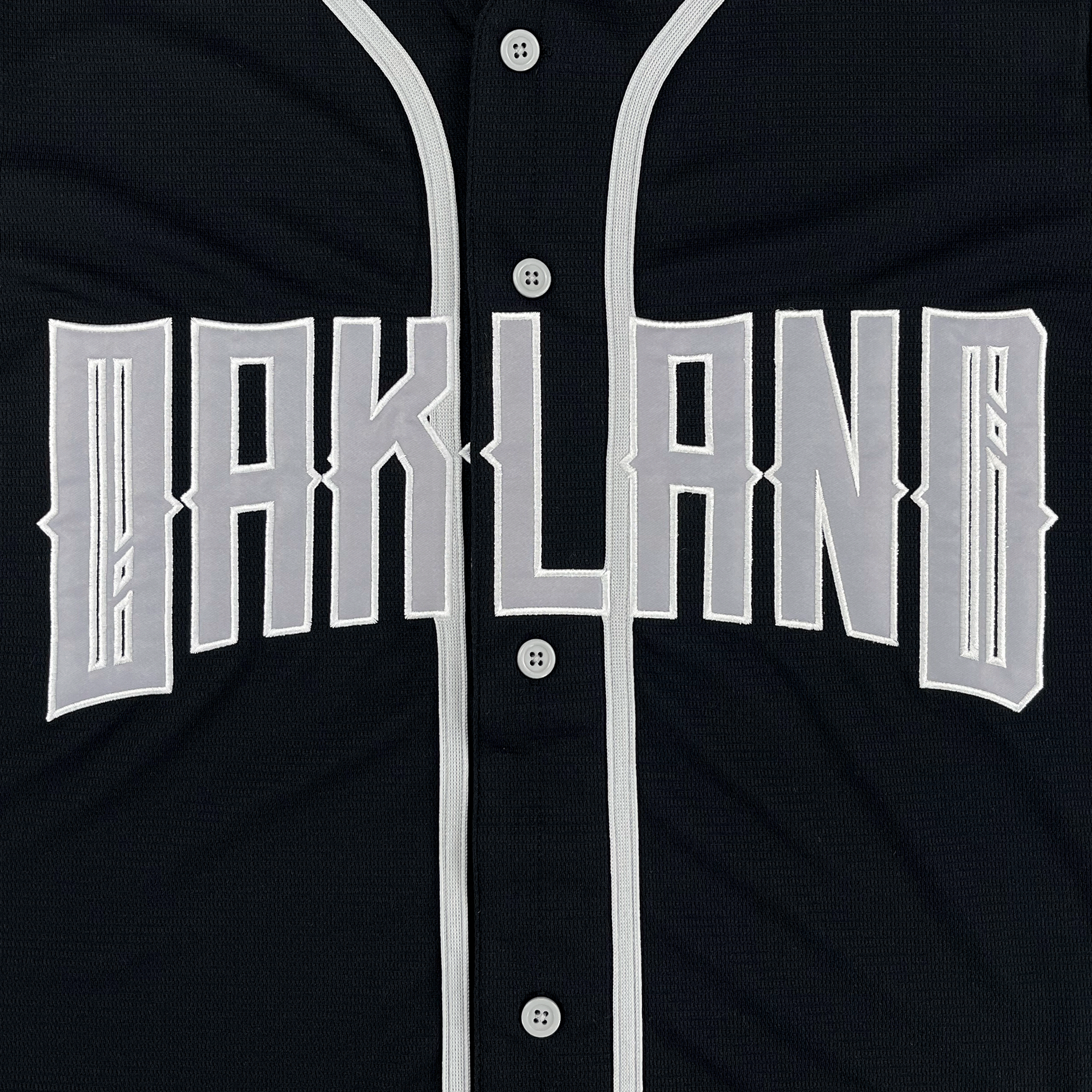 The Oakland Ballers Button Up Jersey by Oakland Ballers is a black jersey with "OAKLAND" in bold white angular letters on the chest, gray vertical piping on the placket and collar, and four gray center buttons.