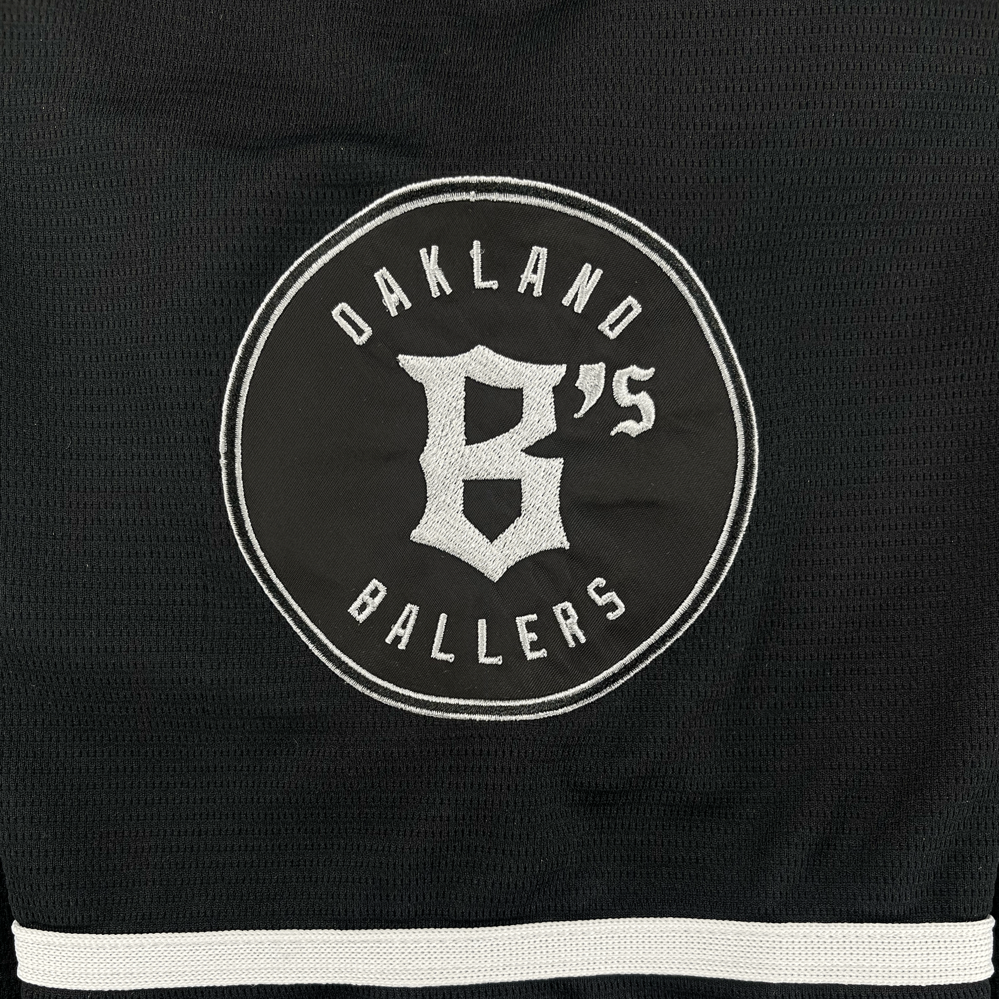 The Oakland Ballers Button Up Jersey by Oakland Ballers features black fabric with a round white embroidered team logo—showing a large “B” and smaller “’S”—and two white stripes along the bottom edge.