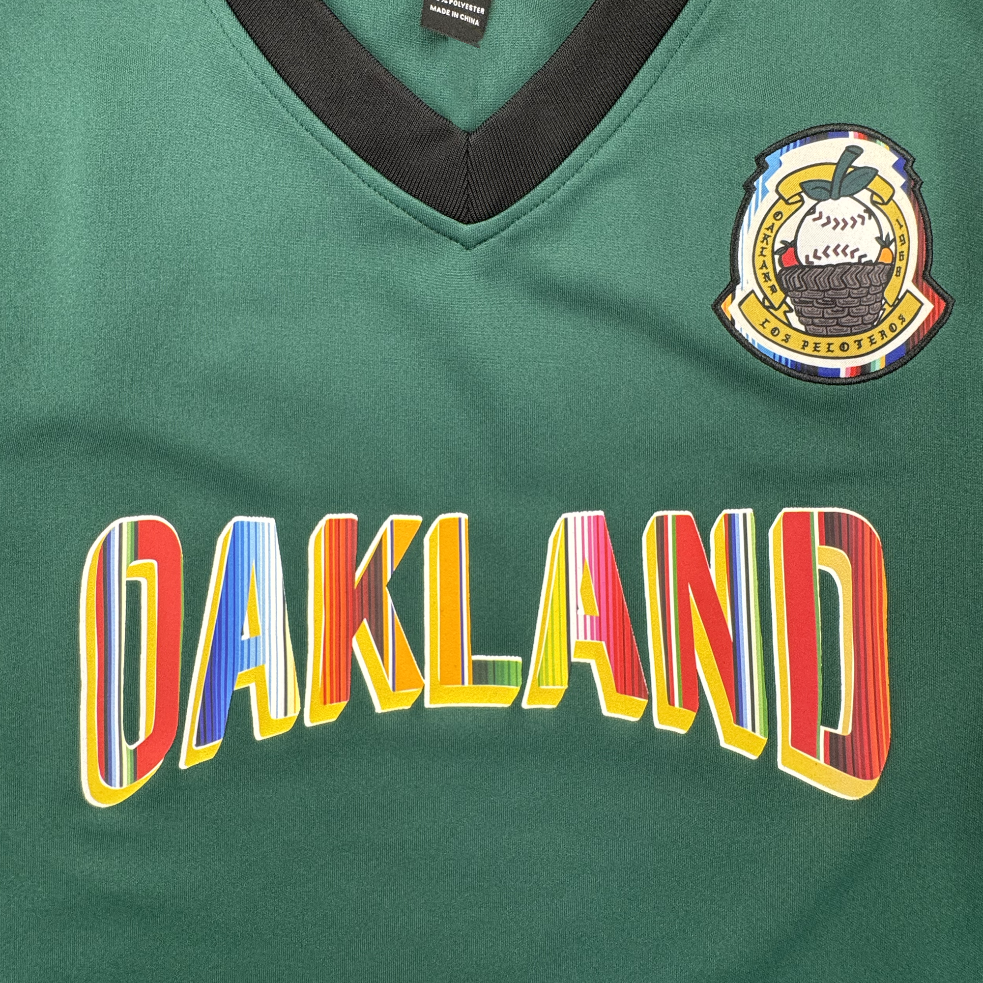 Close-up of a green V-neck jersey with a black collar trim, a multicolor Oakland Peloteros patch on the upper right chest, and large arched 'OAKLAND' lettering across the front in red, yellow, blue, and white.