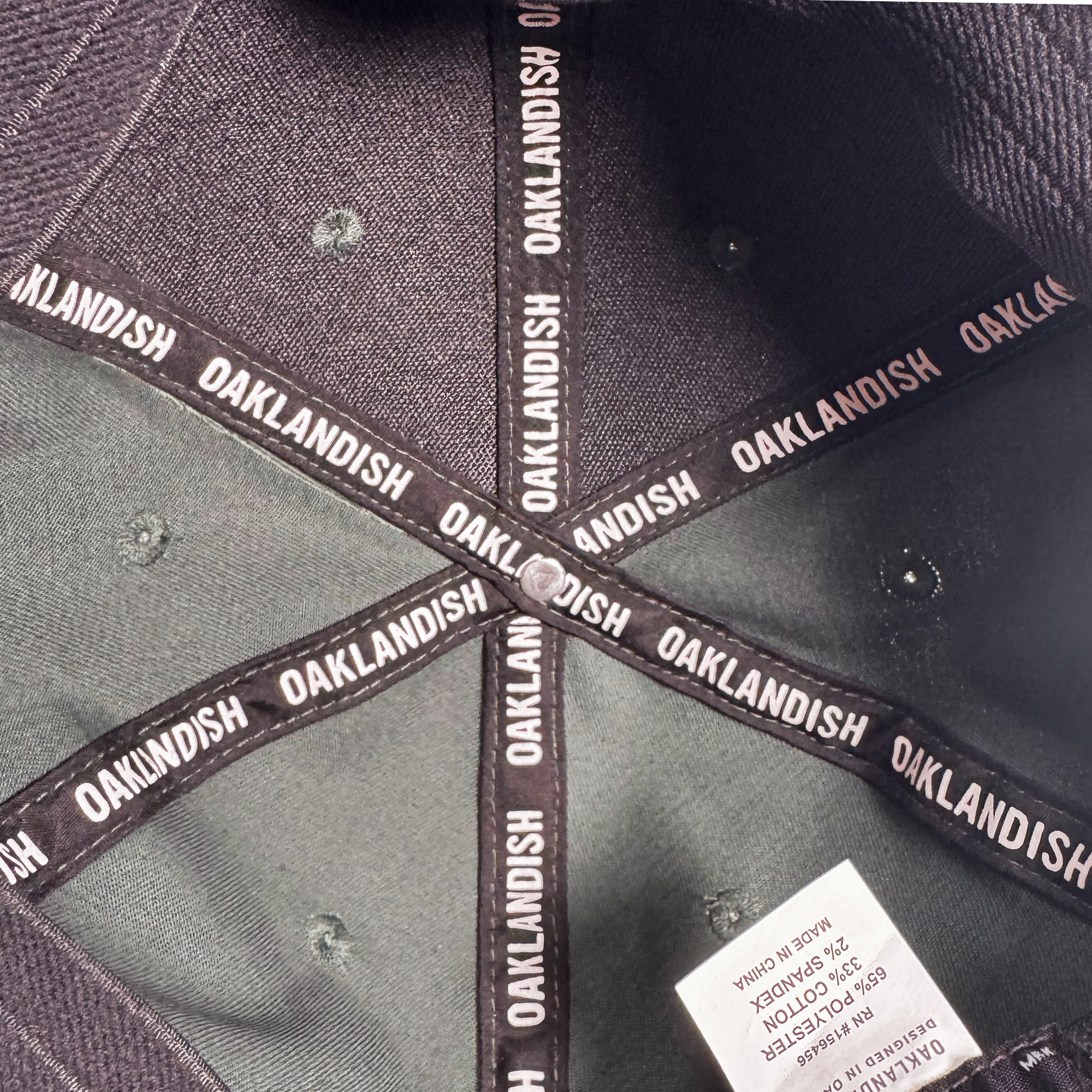Close-up of the Oakland Ballers Script Snapback by Oakland Ballers, showcasing black bands with repeated white "OAKLANDISH" text, a white care tag, visible stitching, and metal ventilation holes inside the black hat.