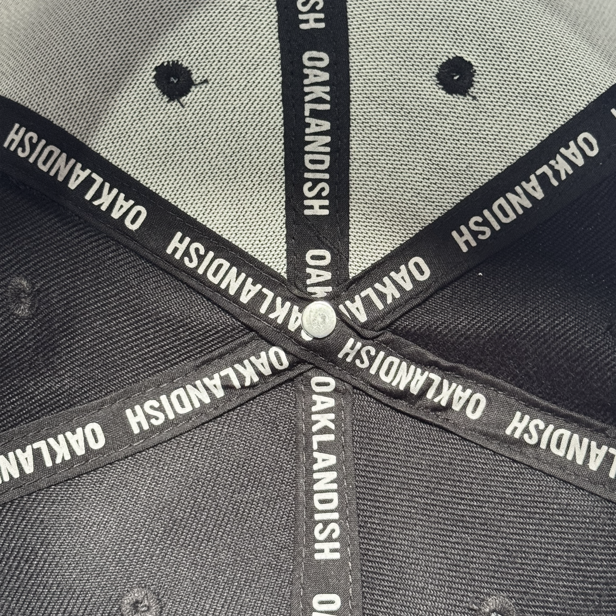 Close-up of the inside of the Oakland Ballers Script Snapback by Oakland Ballers, featuring black fabric strips with repeated white "OAKLANDISH" text forming an X pattern, converging at a silver button, with visible stitching and ventilation holes.