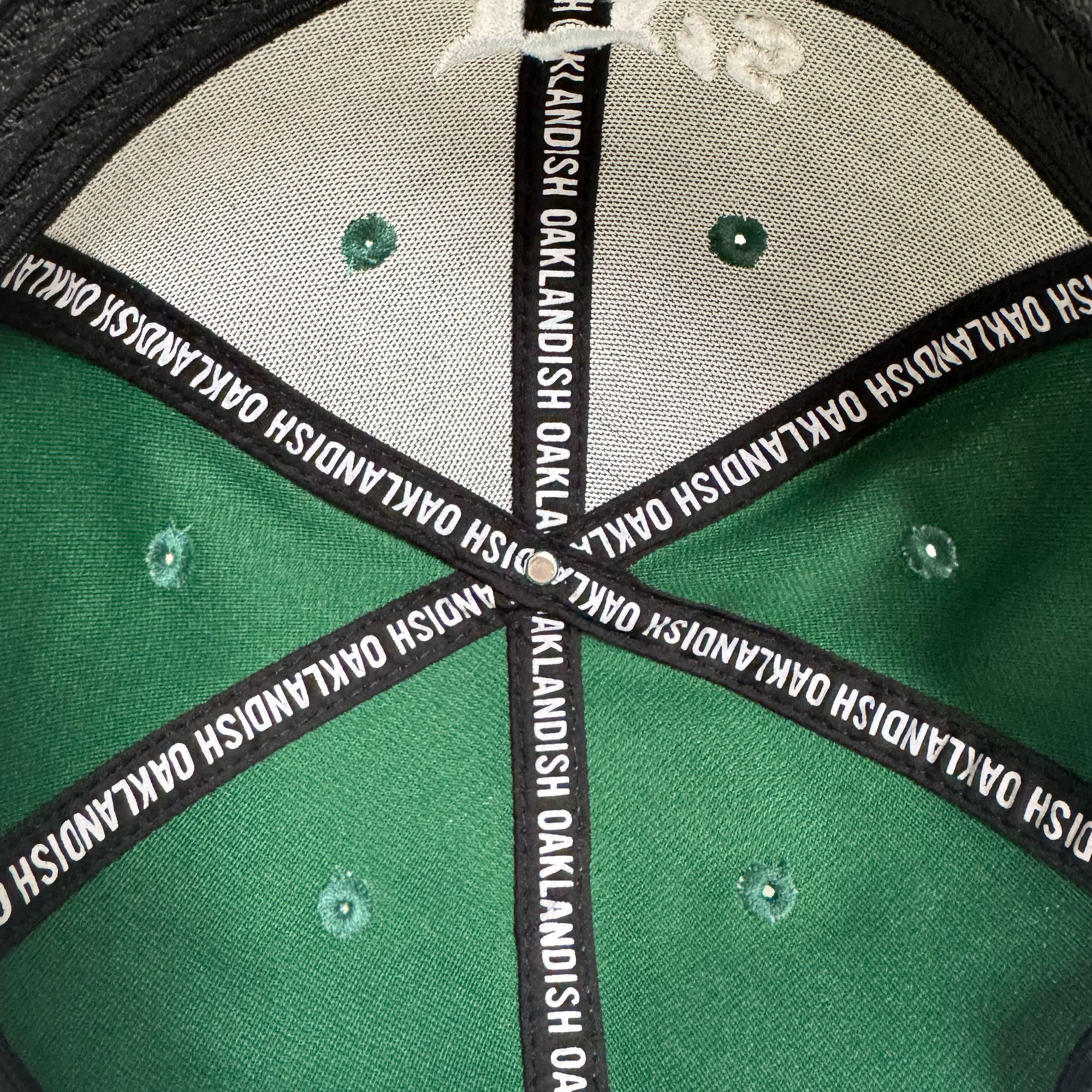 View of the inside top of an Oakland Ballers Logo Flexfit Hat by Oakland Ballers. Black and white taping says "OAKLANDISH" along the seams, with green stitched ventilation holes, celebrating local baseball team pride.