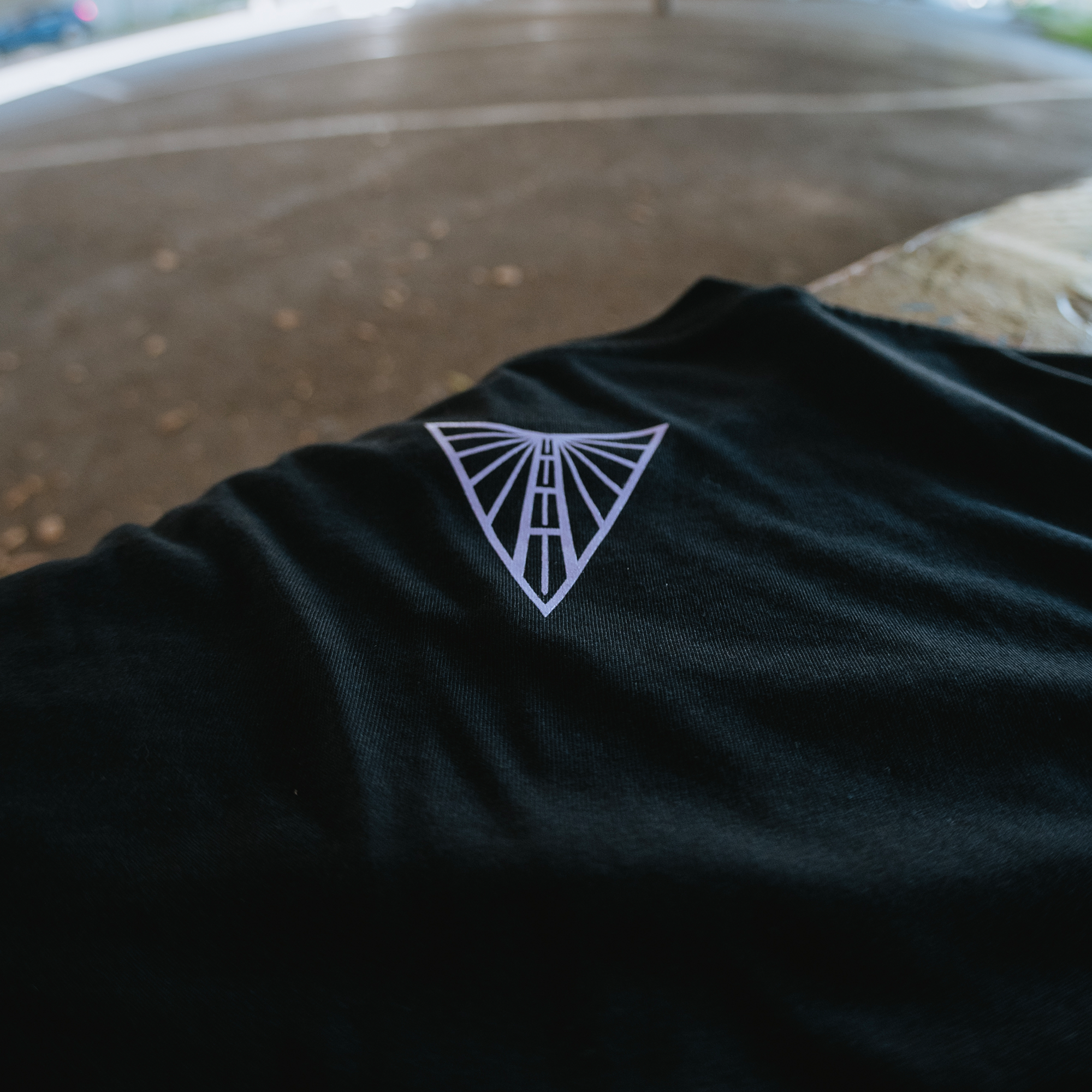 A black cotton Golden State Valkyries Balloons Tee by GSV rests on an outdoor wooden surface, showcasing a white geometric triangle logo. The blurred background displays pavement and greenery—ideal for devoted Valkyries fans.