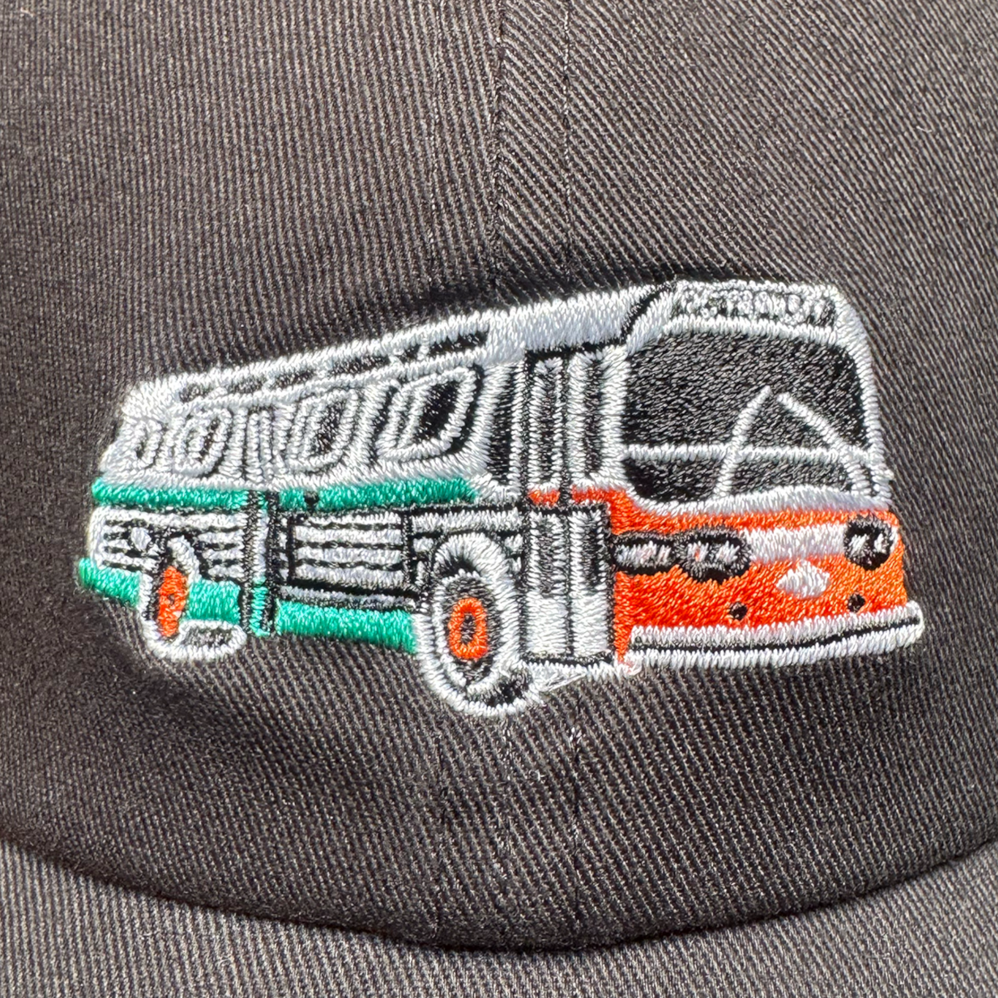 Close-up of the Oaklandish AC Transit Dad Hat's gray fabric, featuring detailed embroidery of a vintage AC Transit bus in orange, green, black, and white thread. The stitching outlines the right-facing bus on the hat's plain gray surface.