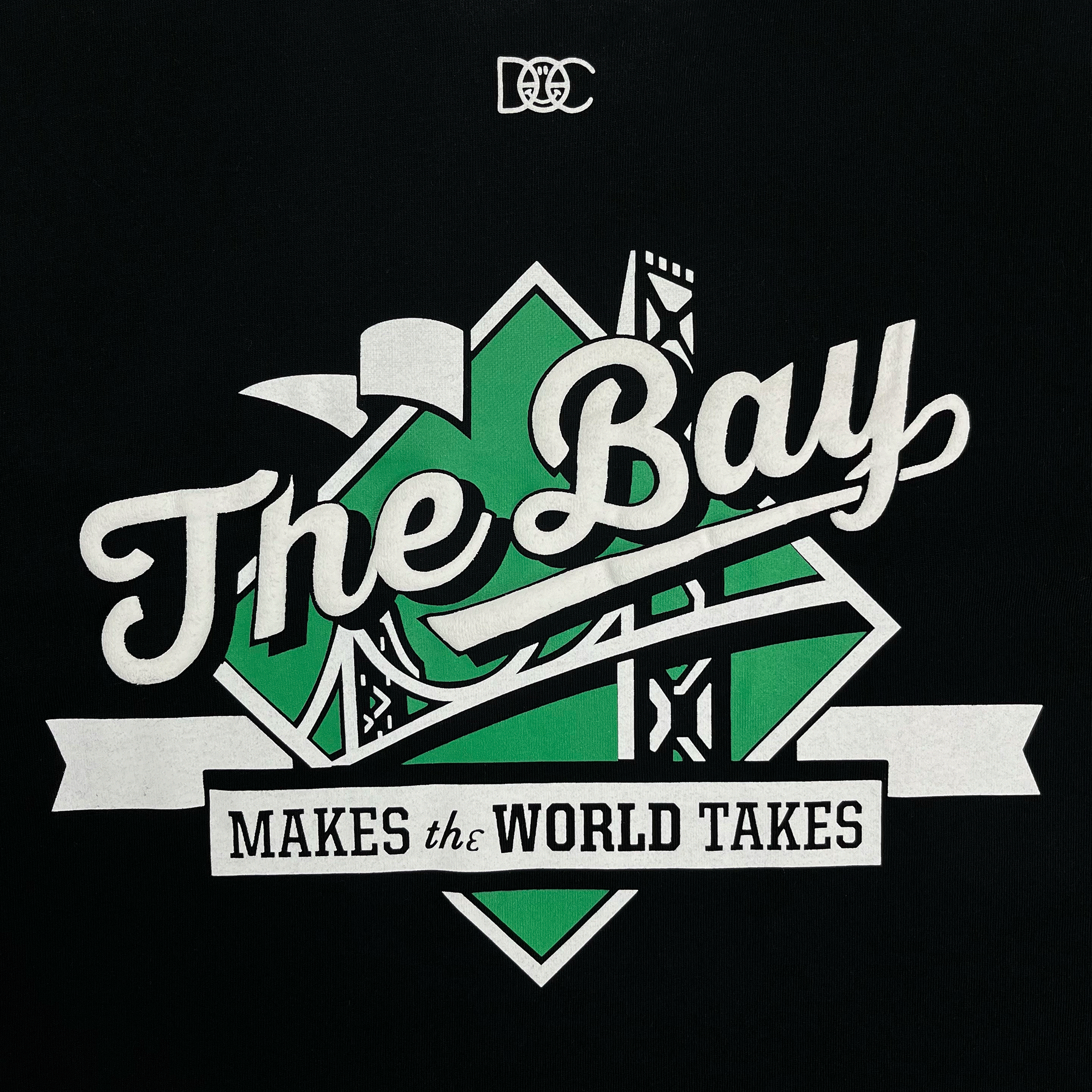 A graphic design on "The Bay Tee by DOC" from Oaklandish showcases the words "The Bay" in large, stylized white script against a green and white geometric background, with ribbon text reading "makes the world takes" below. The entire design is set against a solid black backdrop, featuring a small logo with intertwined letters at the top. This classic fit t-shirt, embodying Bay Area culture in 100% cotton, is perfect for any wardrobe.