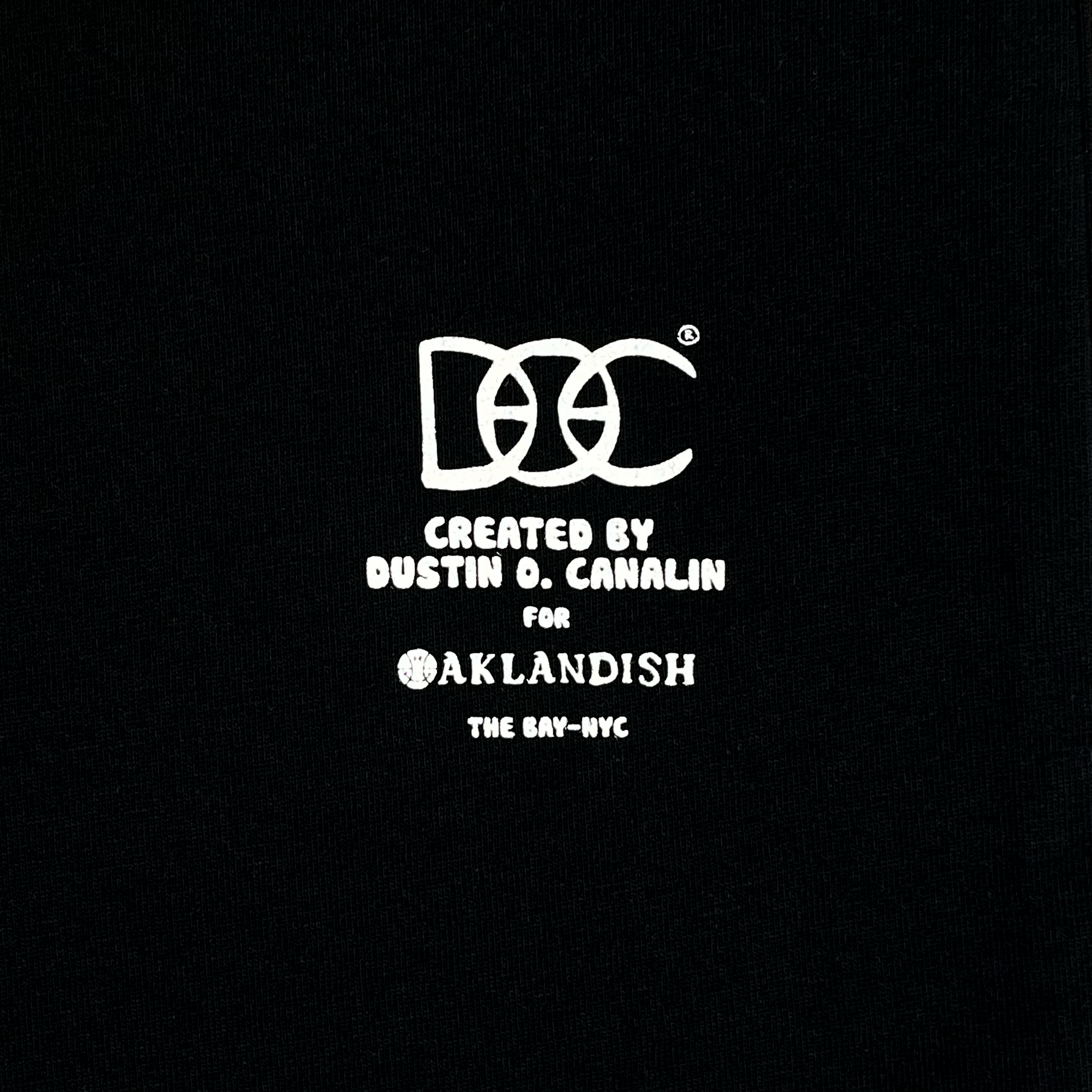 Black background with white text in the center. The text reads "DOZ Created by Dustin O. Canalin for Oaklandish The Bay-NYC." A stylized "DOZ" logo is at the top, with the rest of the text below it, all in a blocky, uppercase font. Celebrating Bay Area culture, this design is also available as The Bay Tee by DOC from Oaklandish in a classic fit t-shirt.