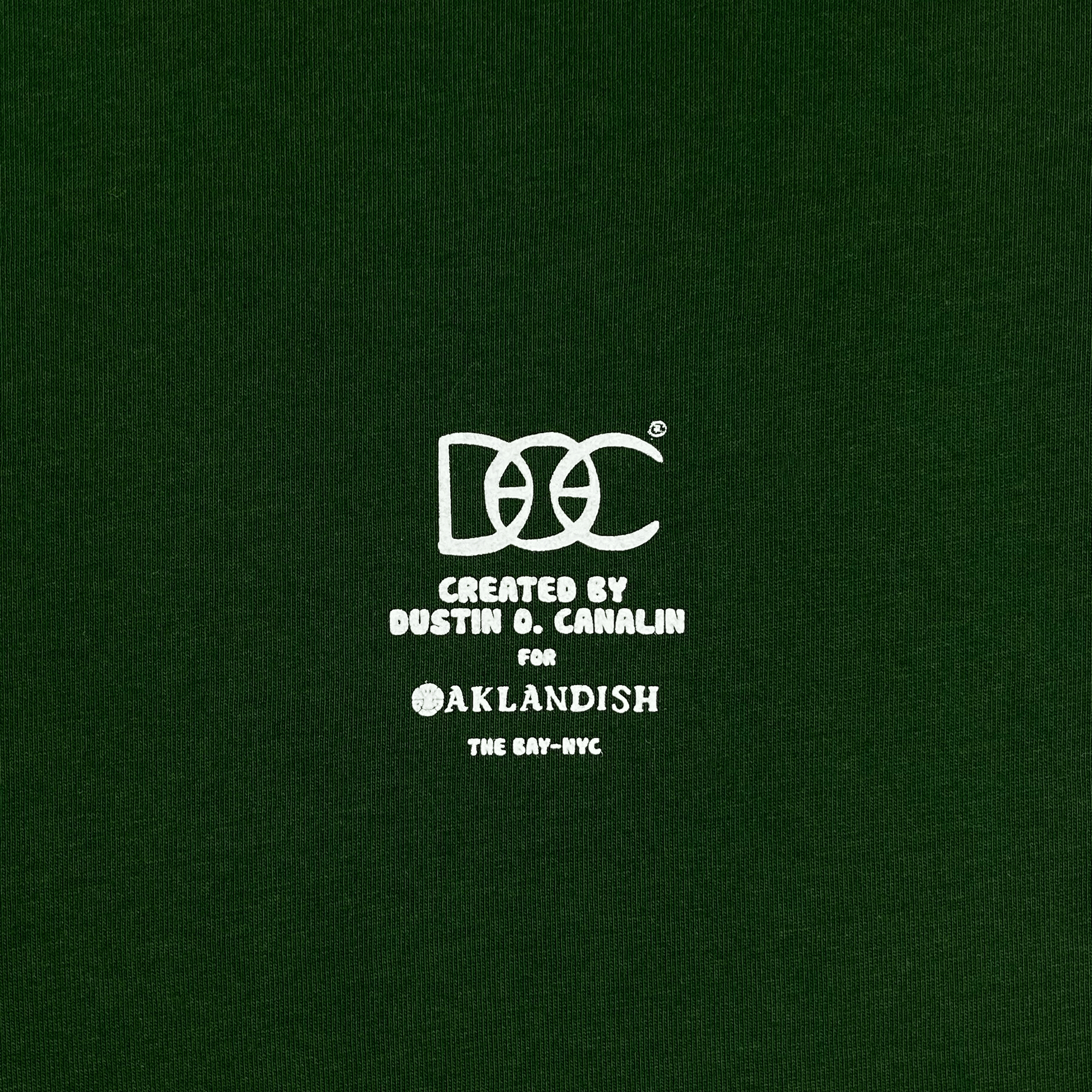 Close-up of white DOC logo Created by Dustin O. Canalin for Oaklandish The Bay-NYC wordmark on a forest green t-shirt.