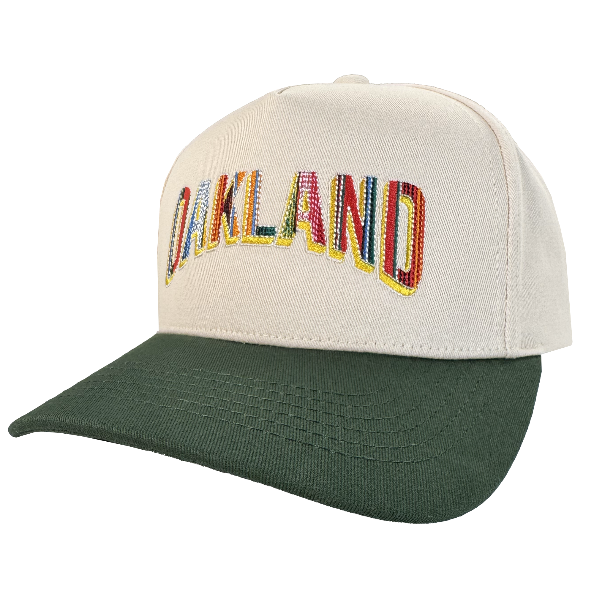 The Oakland Beisbol 5 Panel Cap by Oakland Ballers features a beige pro baseball snapback, dark green brim, and "OAKLAND" in colorful letters across the front—celebrating the team. Displayed angled to show the front panel; white background.