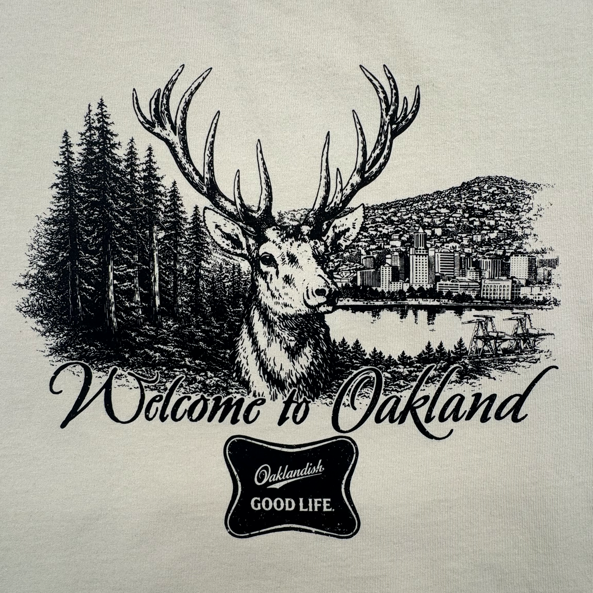 The Oaklandish Buck Tee features a black and white deer with large antlers before pine trees and a cityscape. "Welcome to Oakland" text appears above, with an "Oaklandish GOOD LIFE" badge below. Part of the men's collection.