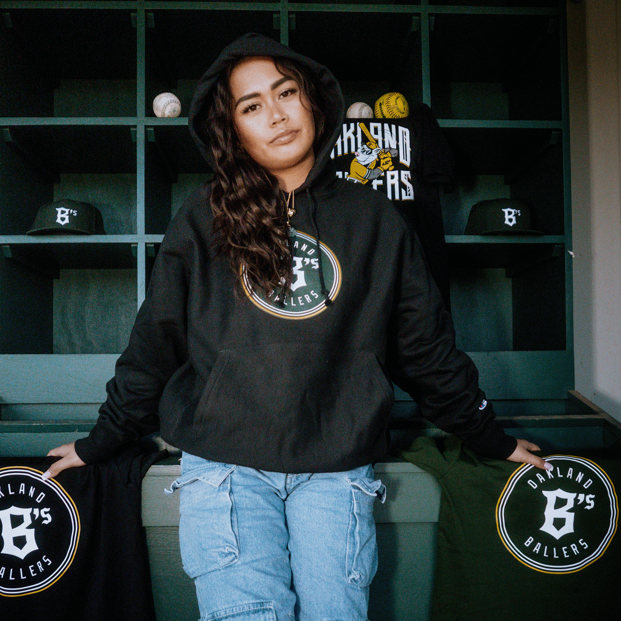 A person with long hair, wearing a Champion X Oakland Ballers Logo Hoodie, stands confidently in front of sports equipment shelves. They are surrounded by Pioneer League gear, including hats and shirts from the Oakland Ballers professional baseball team, in a baseball setting.