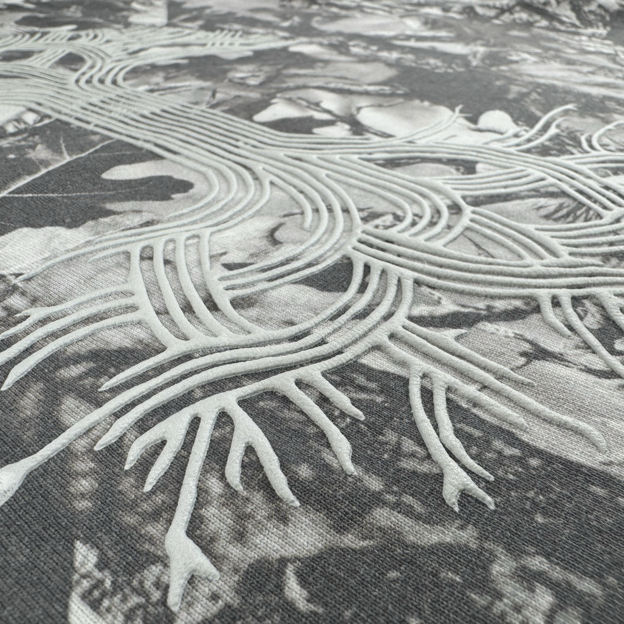 Close-up of raised white embroidery resembling roots on heavyweight cotton with a subtle leafy pattern, sharply contrasting the smooth grey camo background. Shown: Oaklandish Classic Logo Tee by Oaklandish.