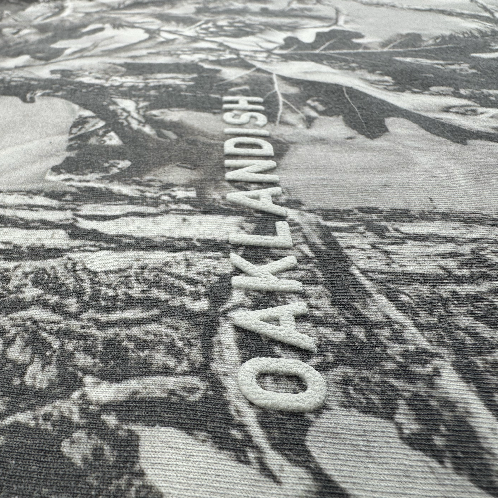A close-up of the Oaklandish Classic Logo Tee by Oaklandish highlights its heavyweight cotton fabric with raised embroidered “OAKLANDISH” lettering over a grey, branch-like camo pattern.