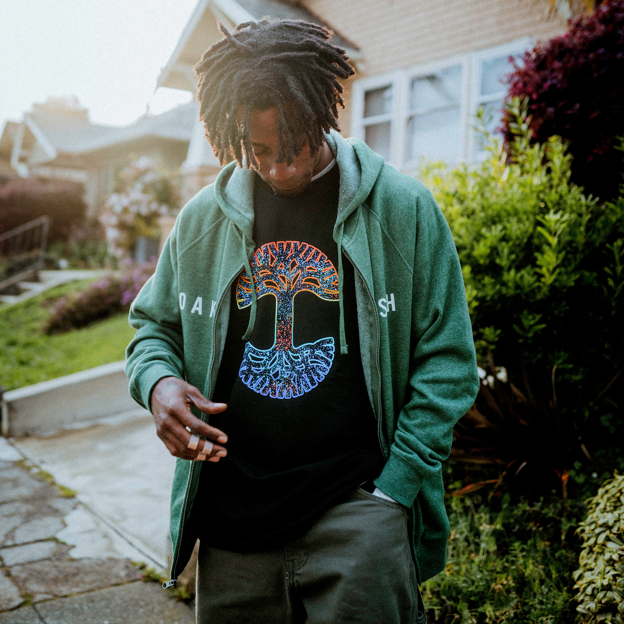 A person with dreadlocks dons a green hoodie and the Oaklandish Prism Tee—a classic fit, 100% cotton shirt with a colorful tree design. They stand on a residential street with houses and greenery, looking down while adjusting their hoodie.