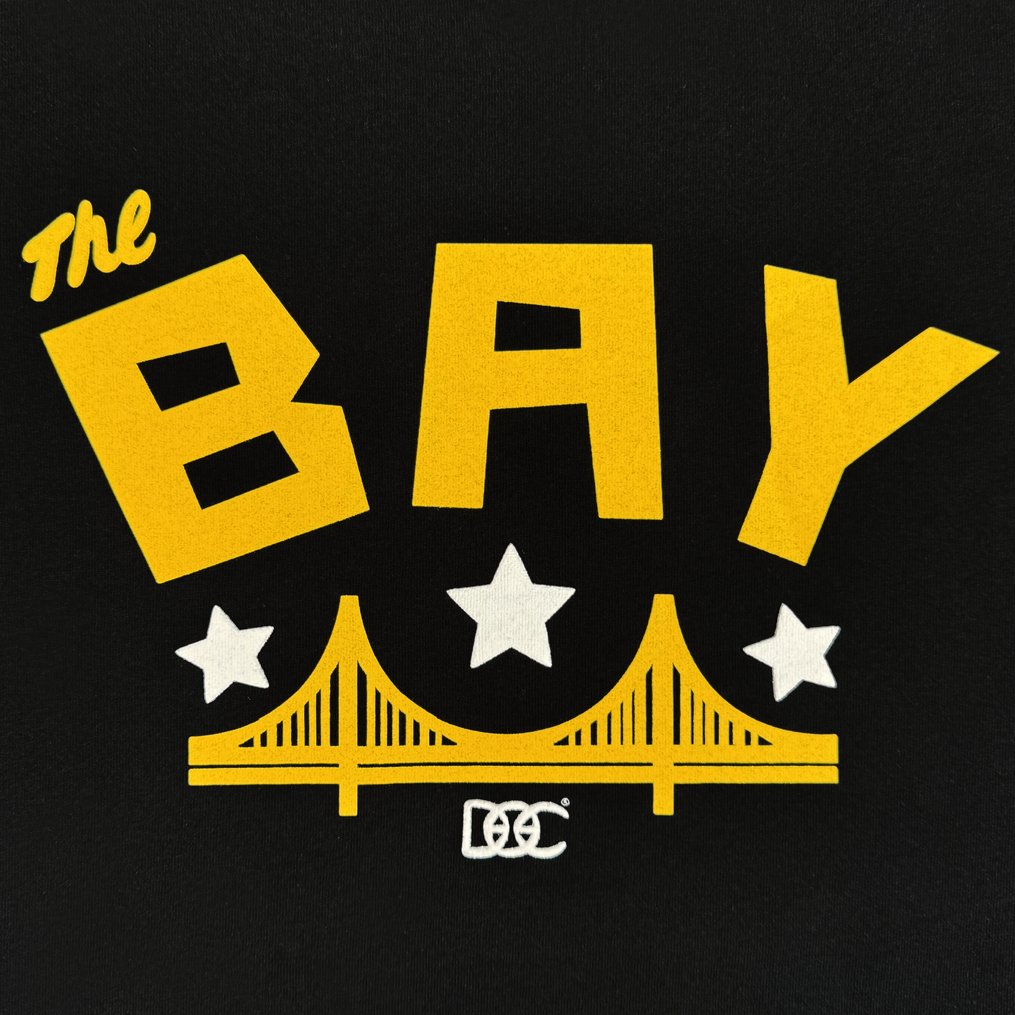 Bold yellow text reading "The Bay" crowns a stylized yellow Golden Gate Bridge and three white stars on black. Below, white overlapping letters "OCC" highlight Oaklandish's Bay Bridge Tee By DOC V2, a classic fit heavyweight cotton shirt.