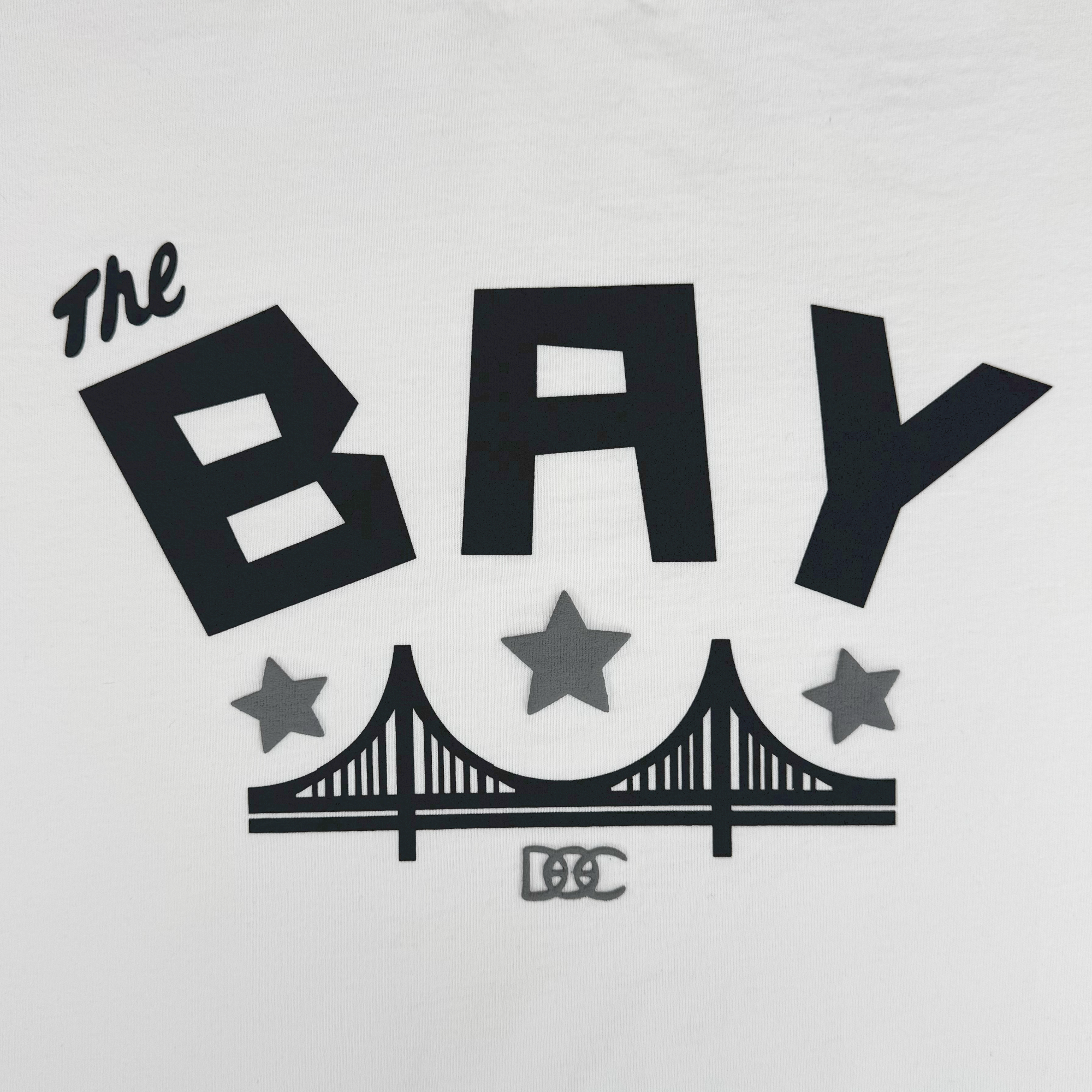 The Bay Bridge Tee By DOC V2 by Oaklandish features bold “The BAY” text, three stars, a stylized bridge graphic, and artistic “DGC” below. Part of The Bay Collection, this classic fit tee is heavyweight cotton in plain white.