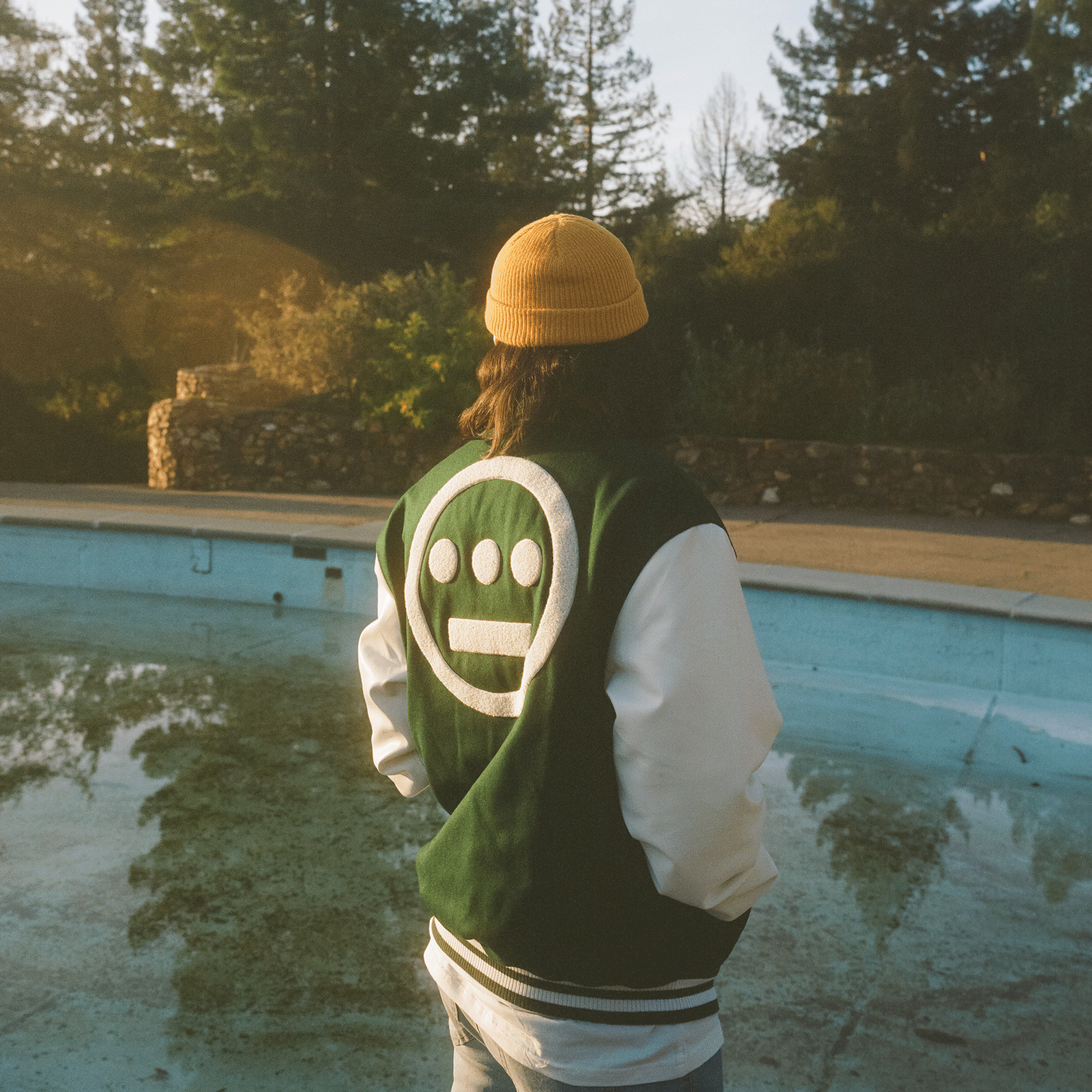 A side view of a man standing beside an abandoned swimming pool, wearing a green varsity jacket with white sleeves and a large white Hieroglyphics hip-hop logo on the center of the back.