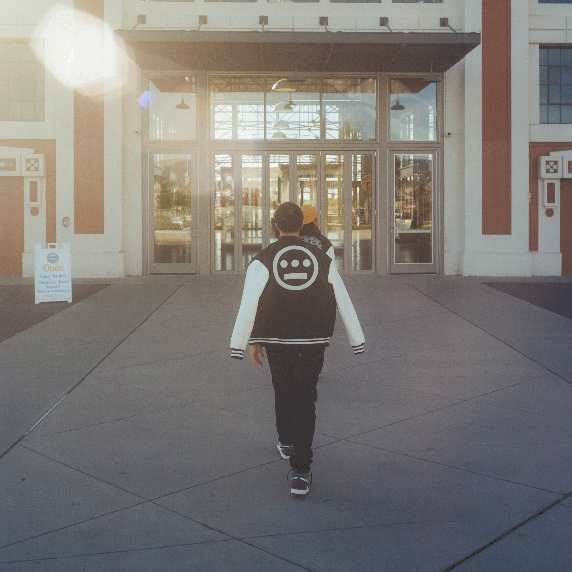 A man walking outdoors, back to the camera, with a green varsity jacket with white sleeves and a large white Hieroglyphics hip-hop logo on the center of the back thrown over his shoulders.
