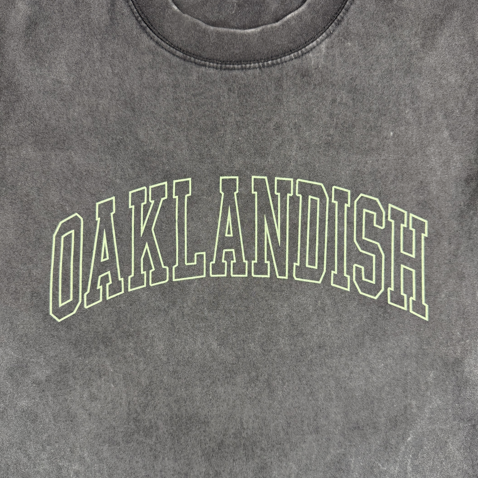 A close-up of the Oaklandish Hunter LS Tee in dark gray, featuring "OAKLANDISH" in large, light green outlined block letters arched across the chest. Made from 100% cotton with a slightly worn, faded texture.