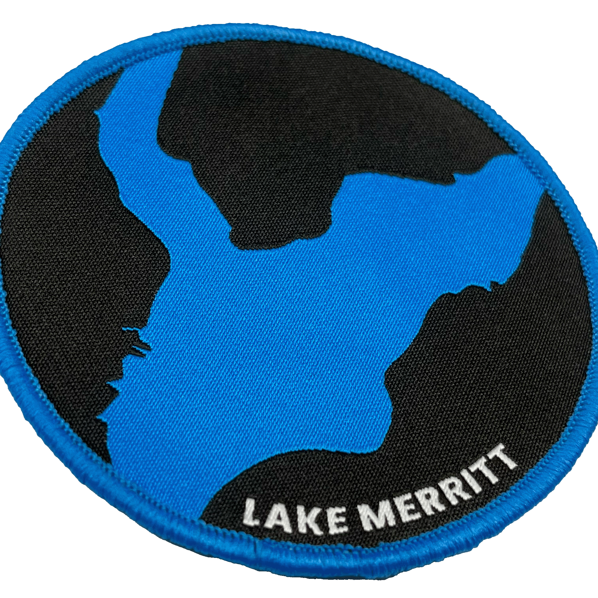 The Oaklandish Lake Merritt Patch features a rounded design with a black background showcasing a blue stylized map of Lake Merritt. The edge is trimmed in blue, and the words "LAKE MERRITT" are elegantly embroidered in white text at the bottom right of this versatile iron-on patch, making it perfect for personalizing your accessories.