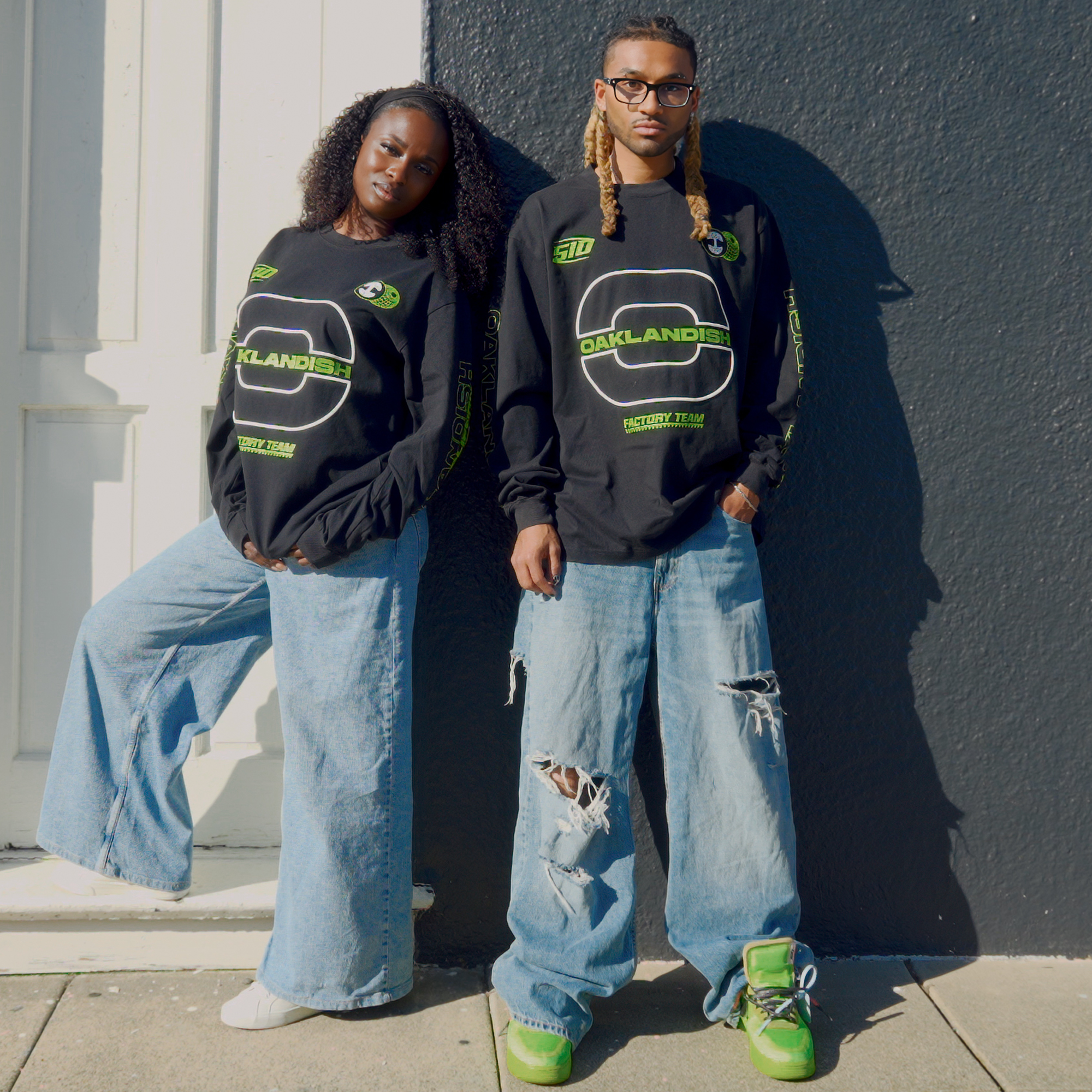 Two people pose by a dark wall, both wearing oversized light blue jeans and black Oaklandish Brrraaap Moto LS Tees. The person on the right pairs theirs with green sneakers and sunglasses for a cool, effortless look.