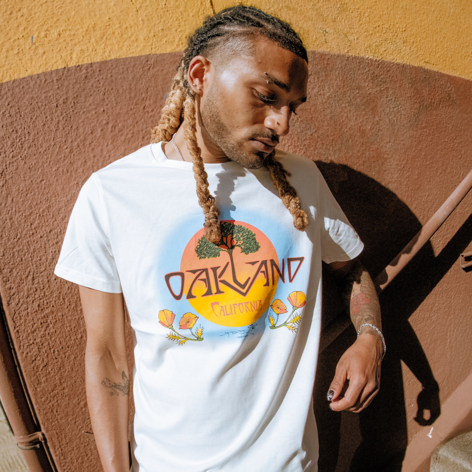 Wearing the Oaklandish Vanguard Tee, featuring "Oakland California" with a sunset, trees, and flowers graphic, a person with long blond-tipped braids stands against a yellow and brown wall, looking down with one hand by their side.