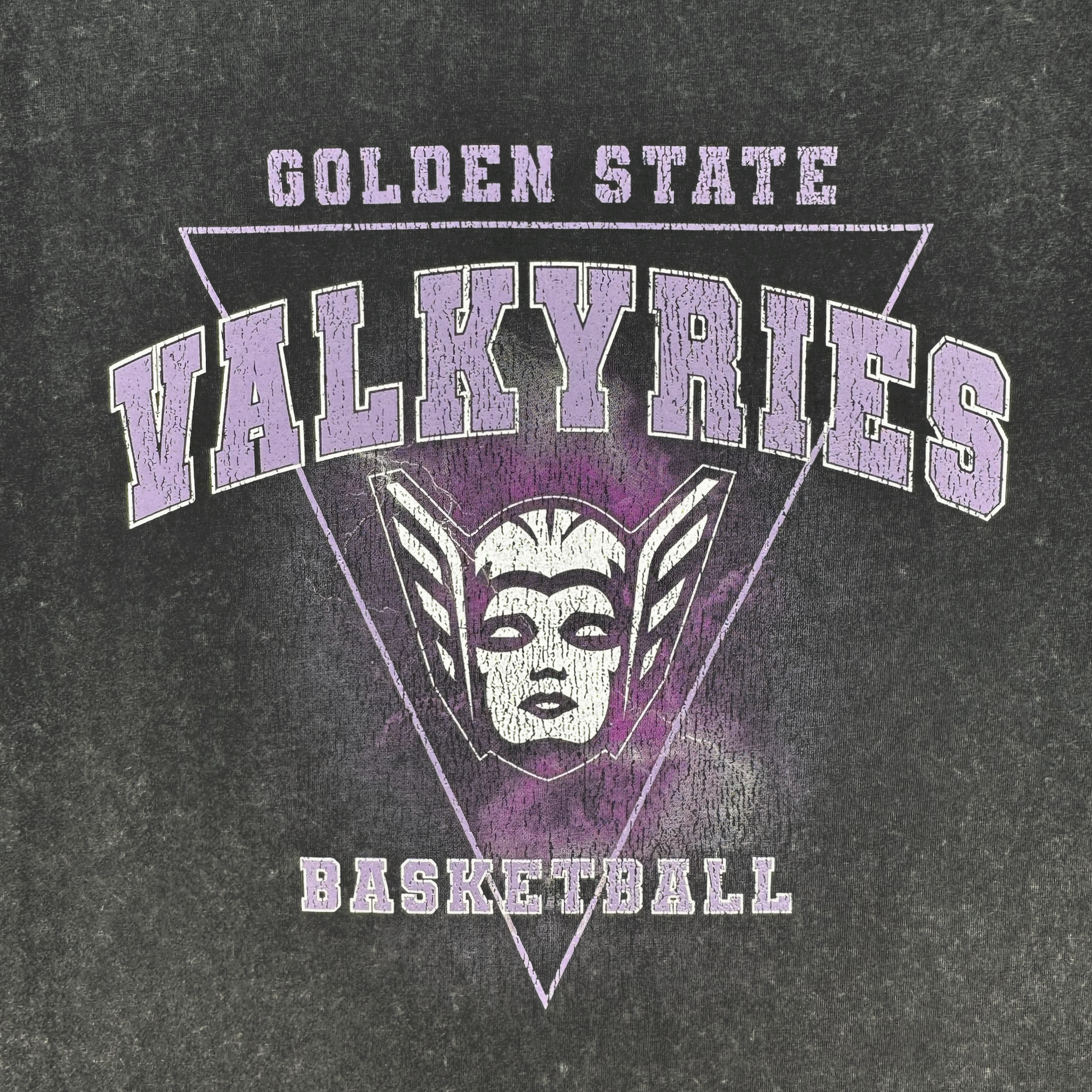 The GSV Golden State Valkyries Surge Stone Wash Heavy Tee features a distressed graphic with a triangle, bold varsity "Golden State Valkyries Basketball" text, and a winged Valkyrie helmet on heavy organic cotton.