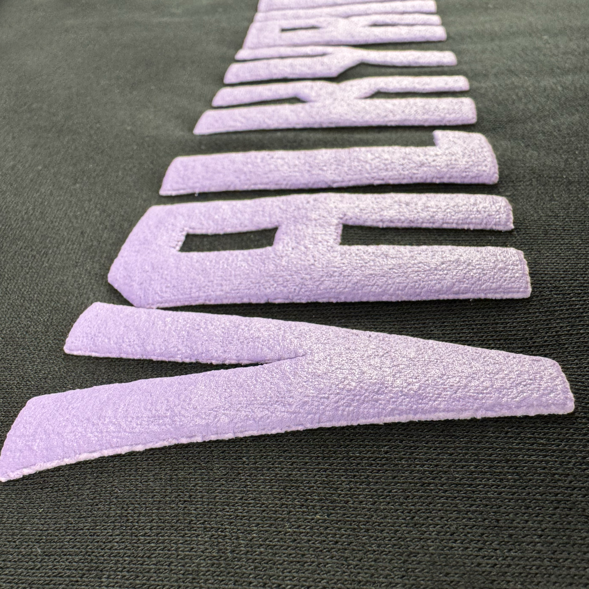 Close-up of textured, raised violet letters spelling "NEW WAVE" diagonally on a black mid-weight fleece fabric. The soft, suede-like text stands out against the smooth material of the GSV Golden State Valkyries Violet Lightning Hoodie.