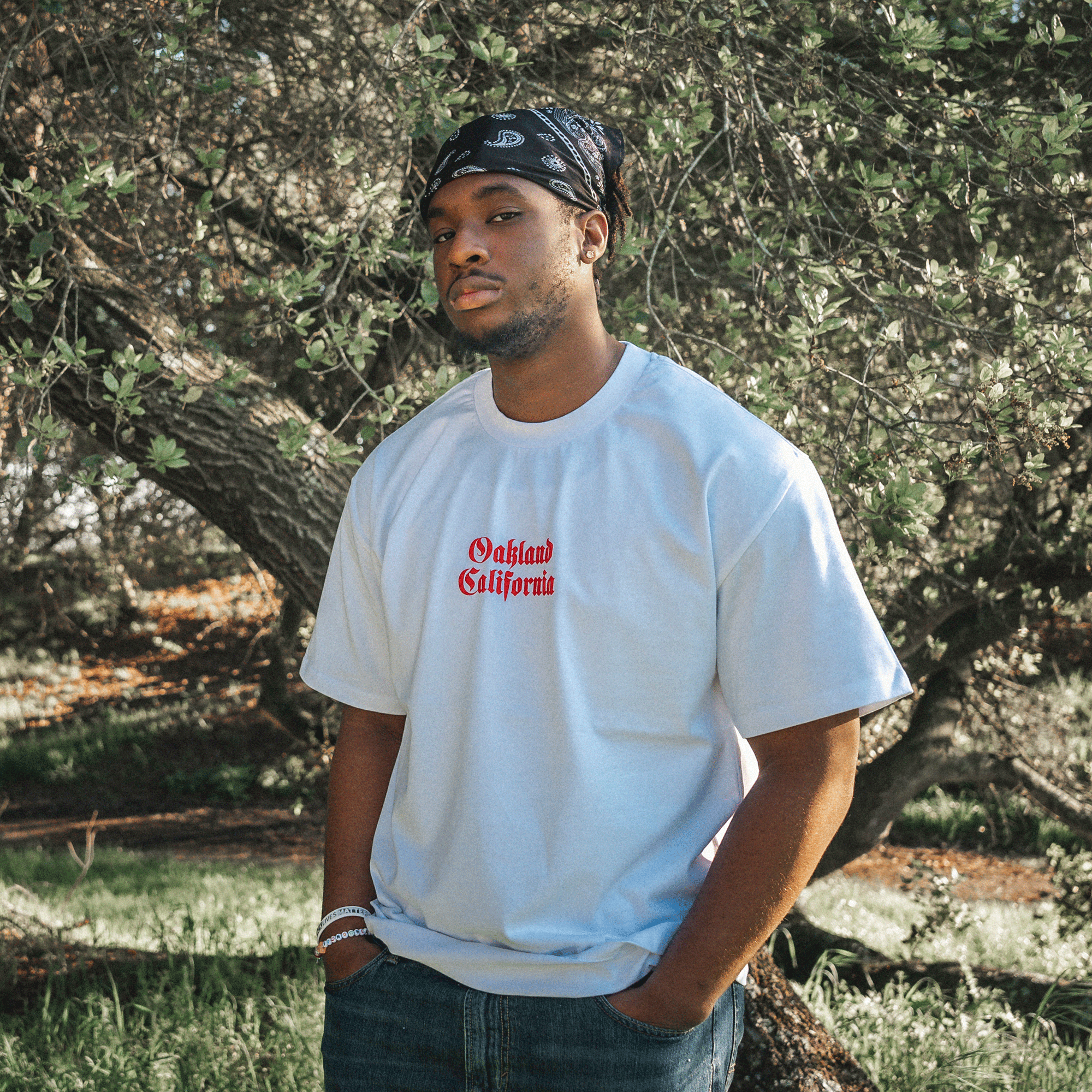 A man stands outdoors in front of a tree, wearing a white Town Rose Heavy Tee by Oaklandish with red text that reads "Oakland California." The men's sizing t-shirt has a boxy fit. He also sports a black bandana on his head and blue jeans. The background showcases lush greenery and a sunlit forest area, while his expression remains neutral.