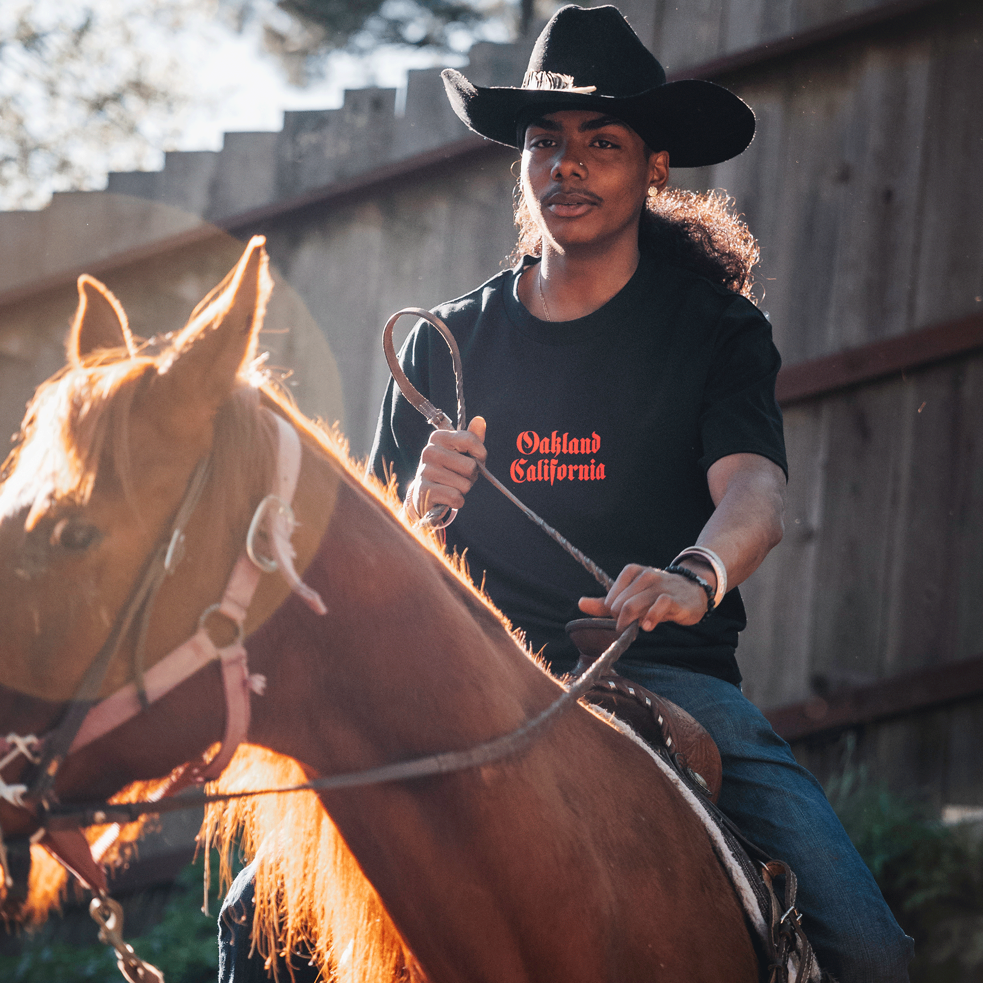 Model on a horse wearing black tee with Oakland California printed centered in red ink.