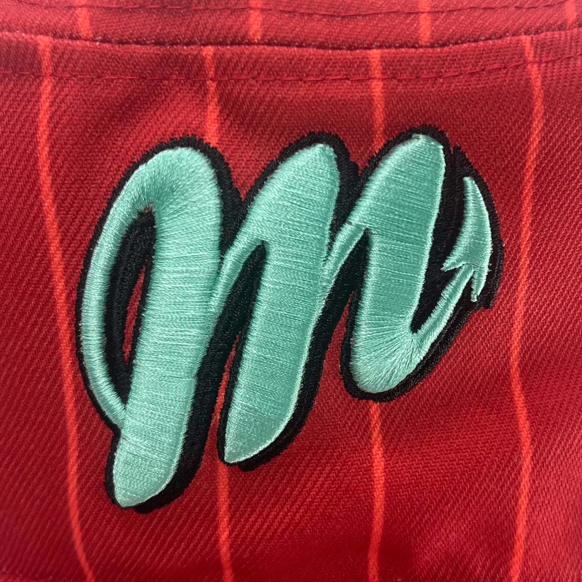 A close-up view of an embroidered logo on a New Era Diablos Paisley Bucket hat by Retail Vendors, featuring a light blue letter "M" with a black outline, resembling a stylized brushstroke. The letter has a decorative flourish that turns into an arrowhead. The background is red polyester fabric with subtle vertical stripe patterns visible.